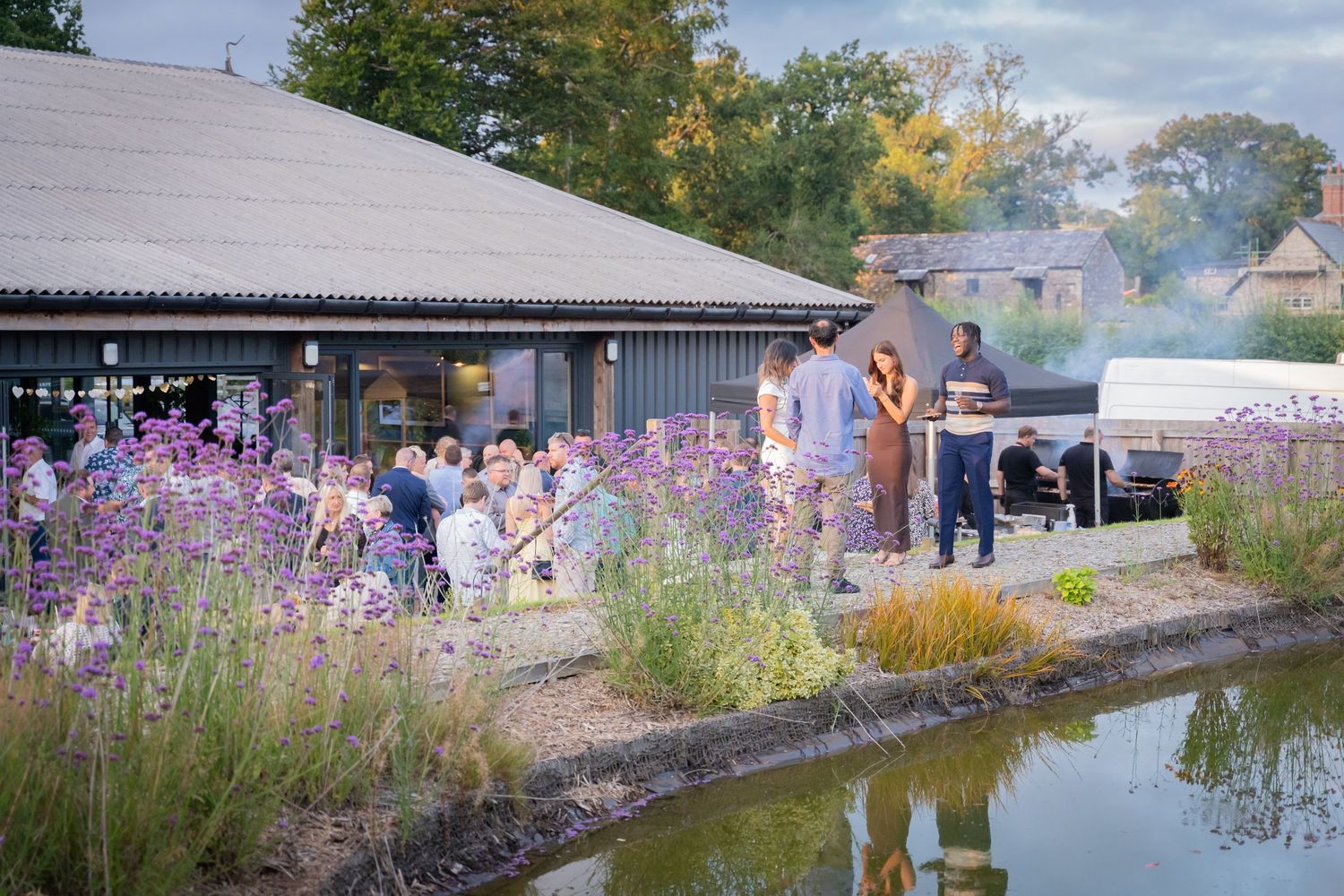 A serene waterfront walkway with wildflowers and native plants along a tranquil pond on a cloudy autumn day.