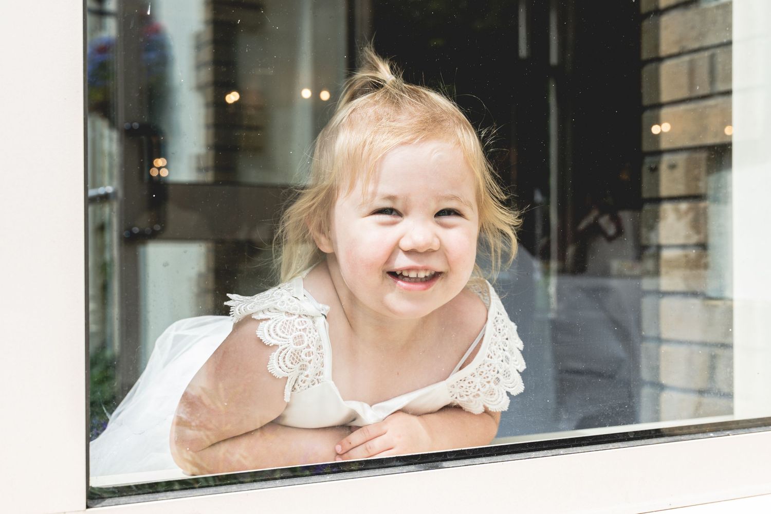 A young child leans on a windowsill and smiles while wearing a white lace-trimmed dress.