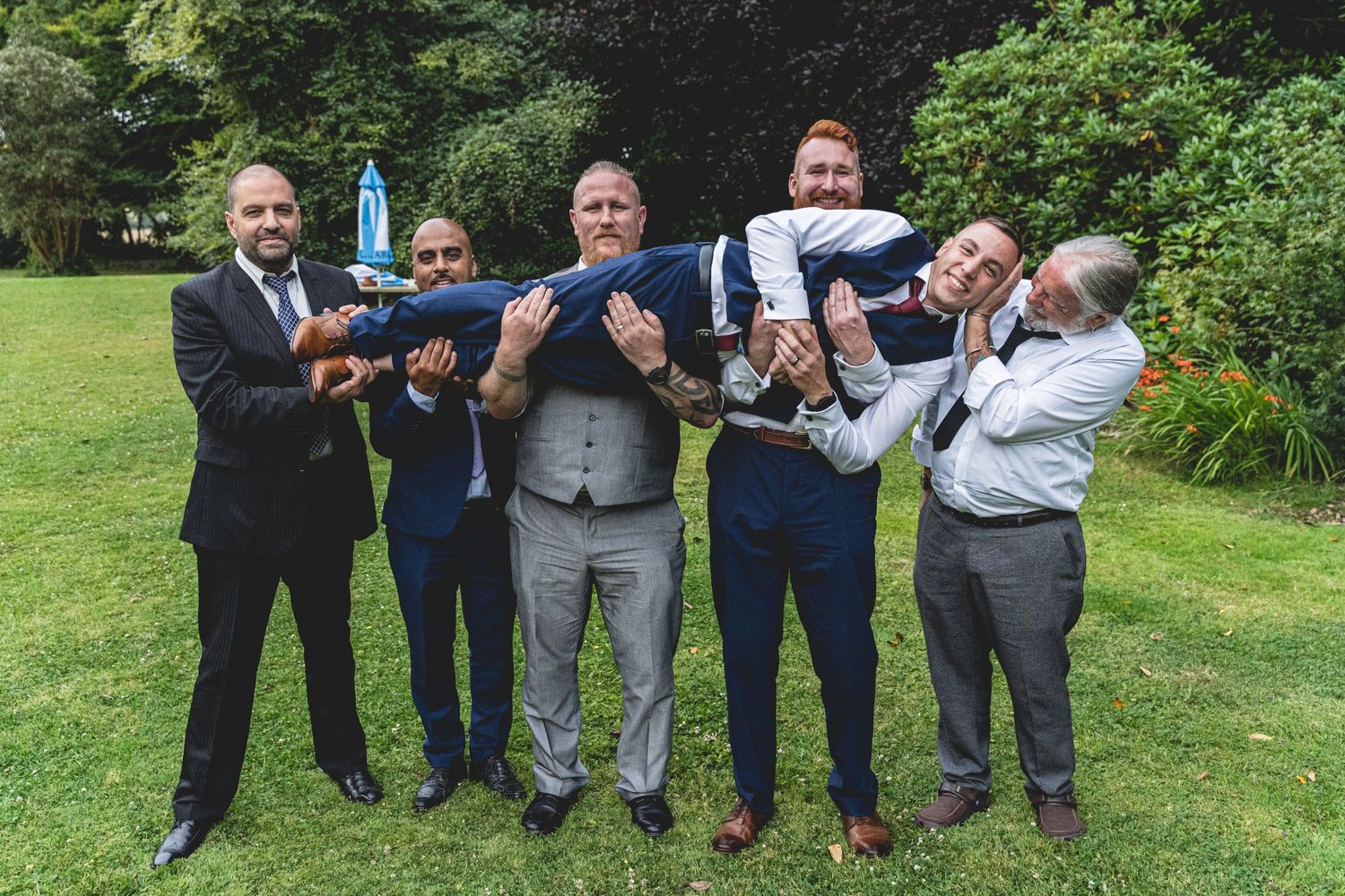 Groomsmen lifting someone in playful pose on garden lawn. Cornwall Wedding Photographer. Groomsmen Cornwall.
