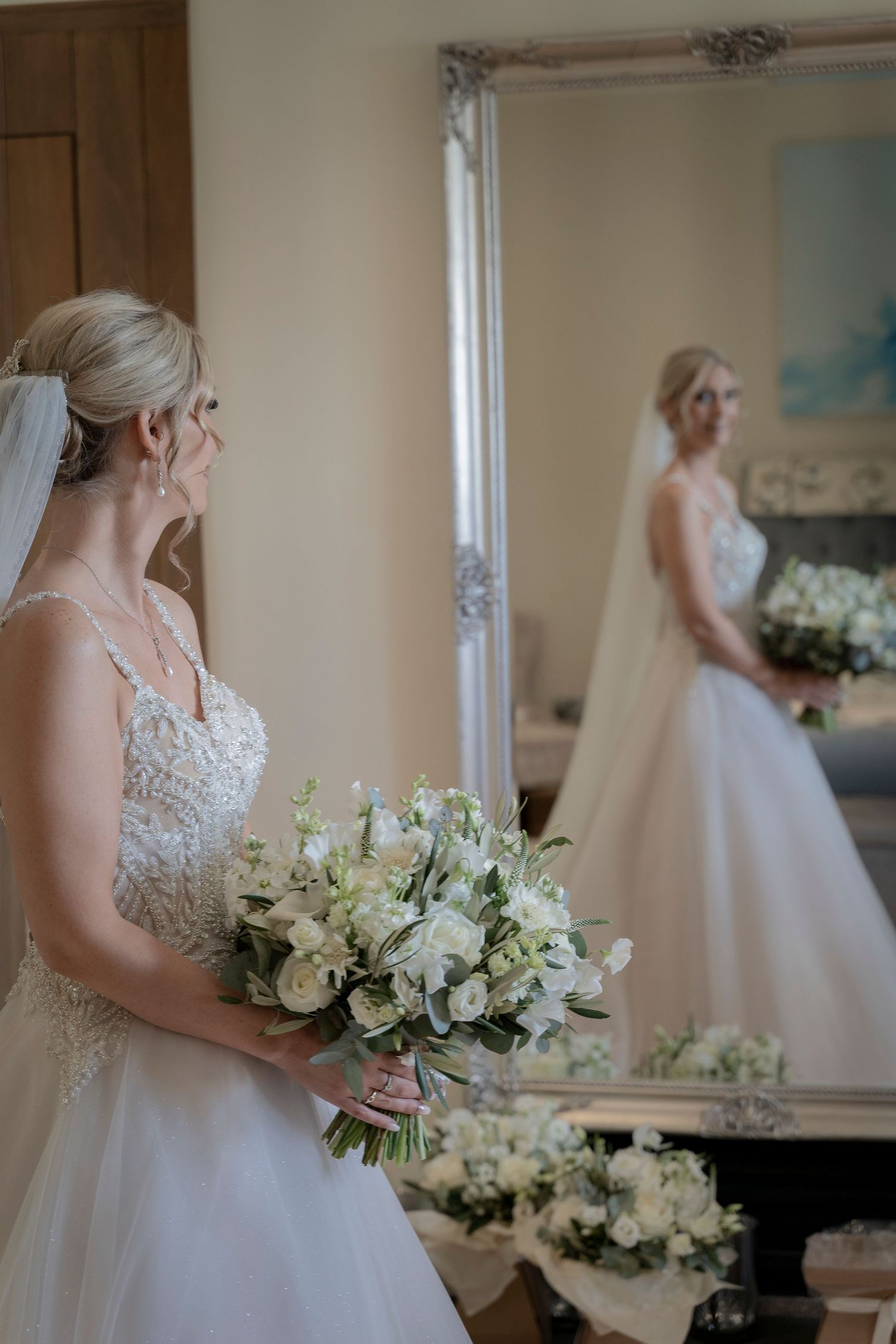 A bride in a lace wedding gown holds a white bouquet while looking in an ornate mirror.