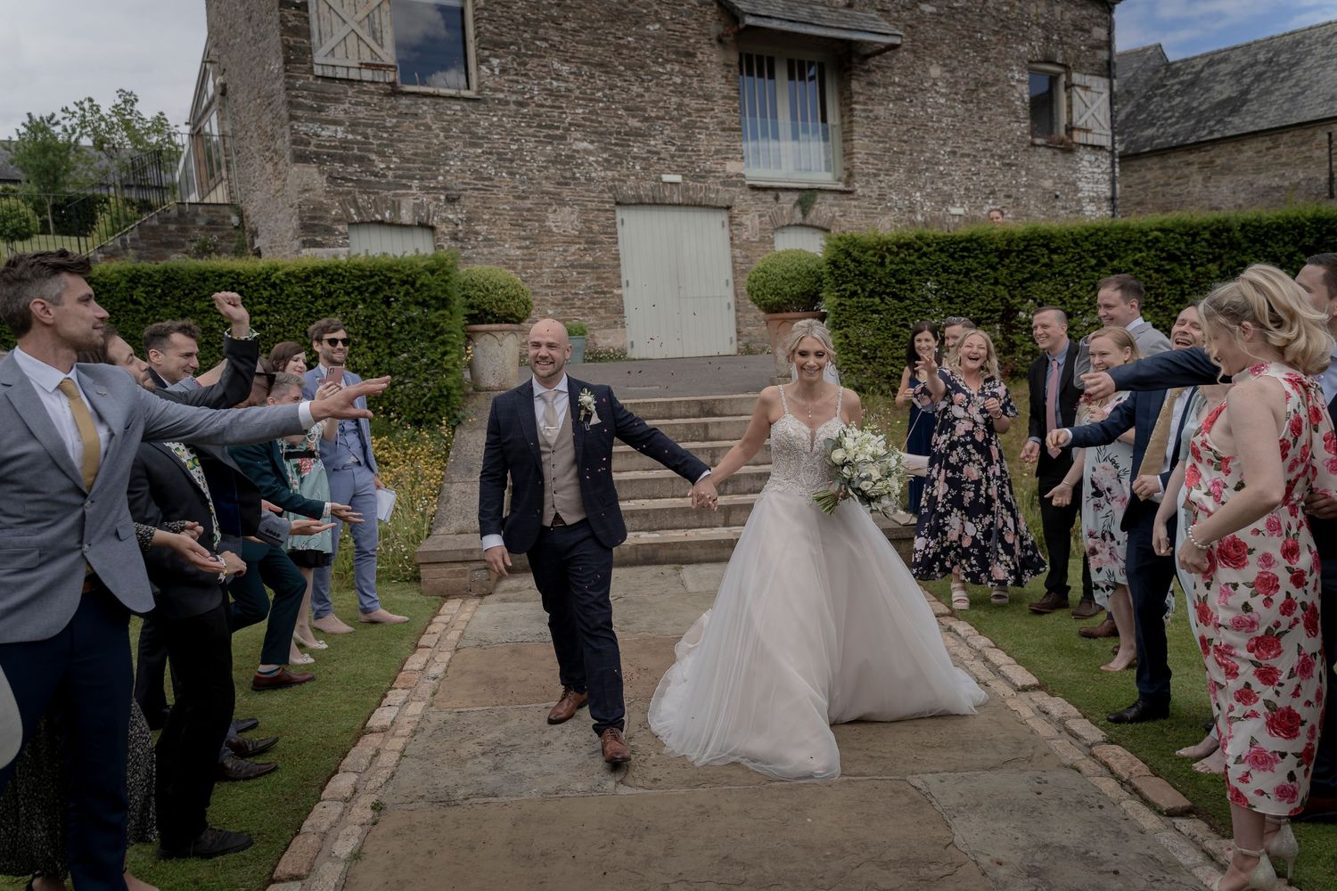 Wedding guests form an aisle outdoors as newlyweds exit stone building celebrations. Shilstone House Wedding. Devon Wedding Photography.