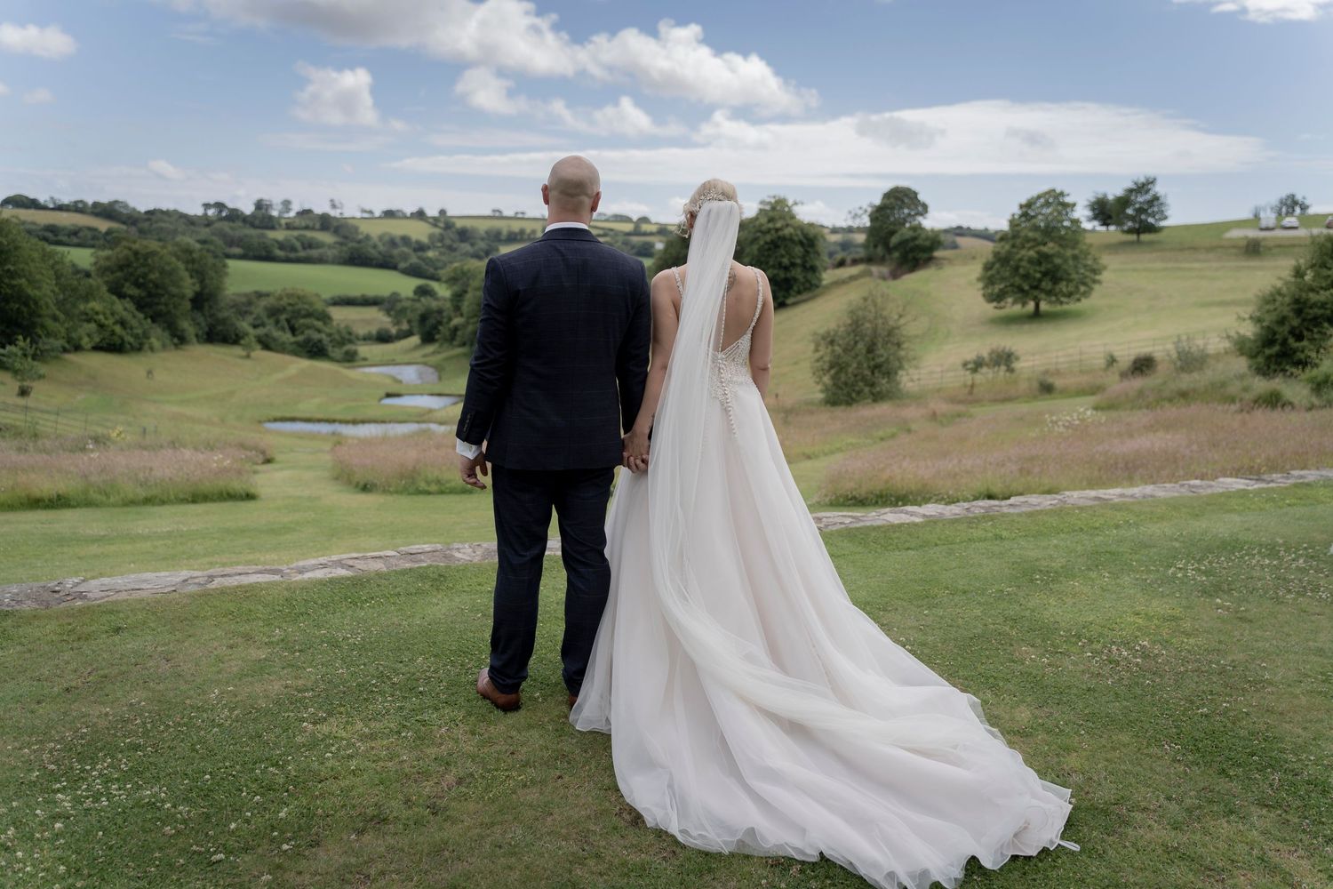 Couple stands at hillside overlook with flowing wedding dress and countryside views. Shilstone House, Devon Wedding. Devon Photographer.