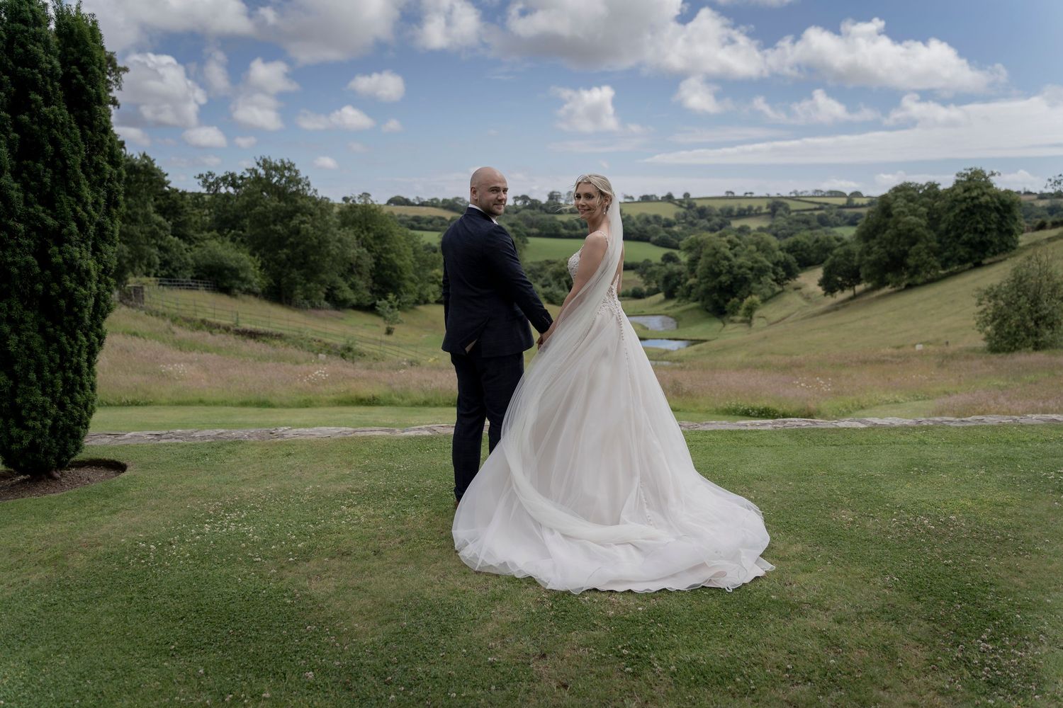 Newlyweds pose together on hilltop with sweeping valley views on sunny day. Shilstone House, Devon Wedding. Devon Photographer.