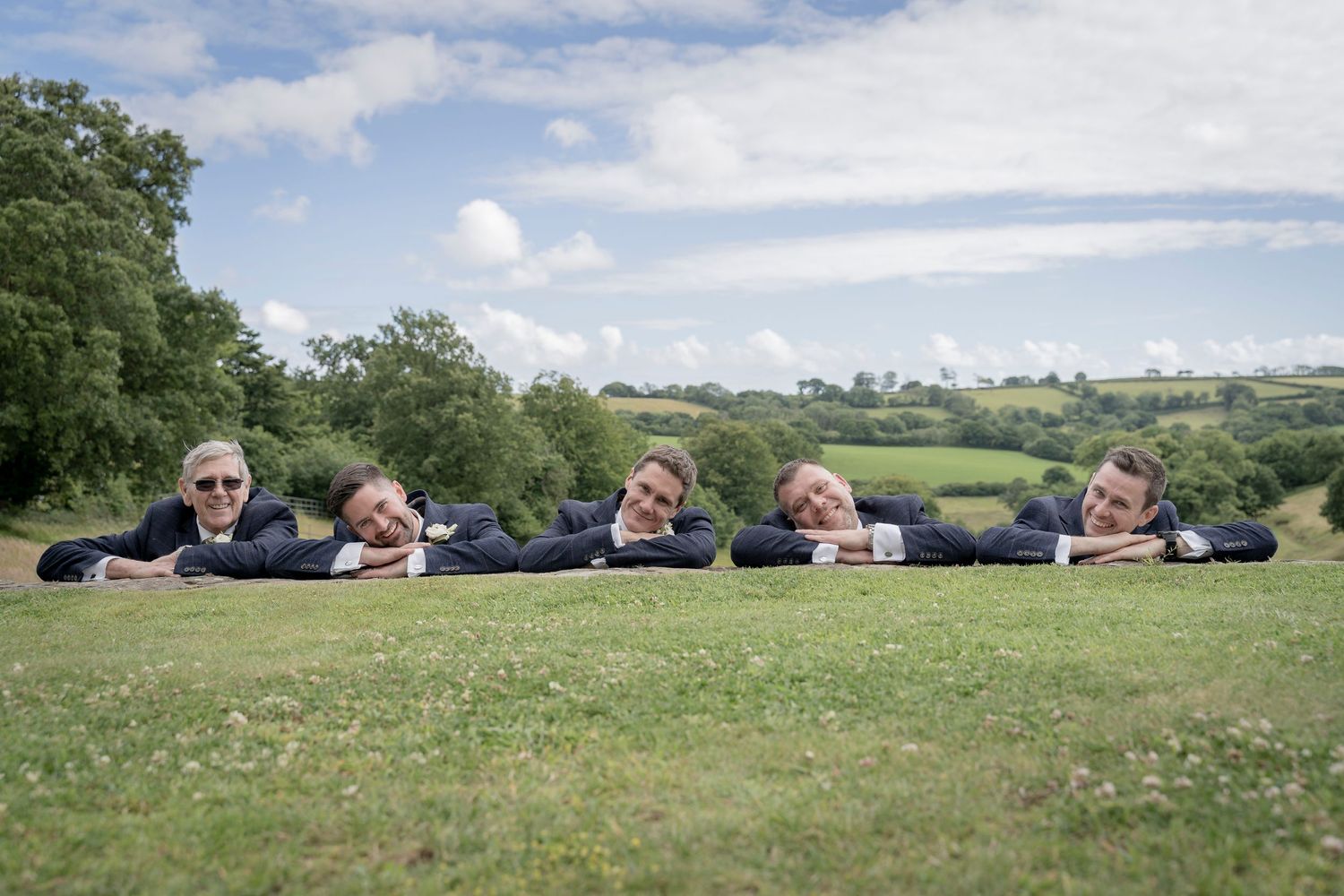 Group lying in a line on green grass during a fun wedding photoshoot in countryside setting.   Groomsmen posing.