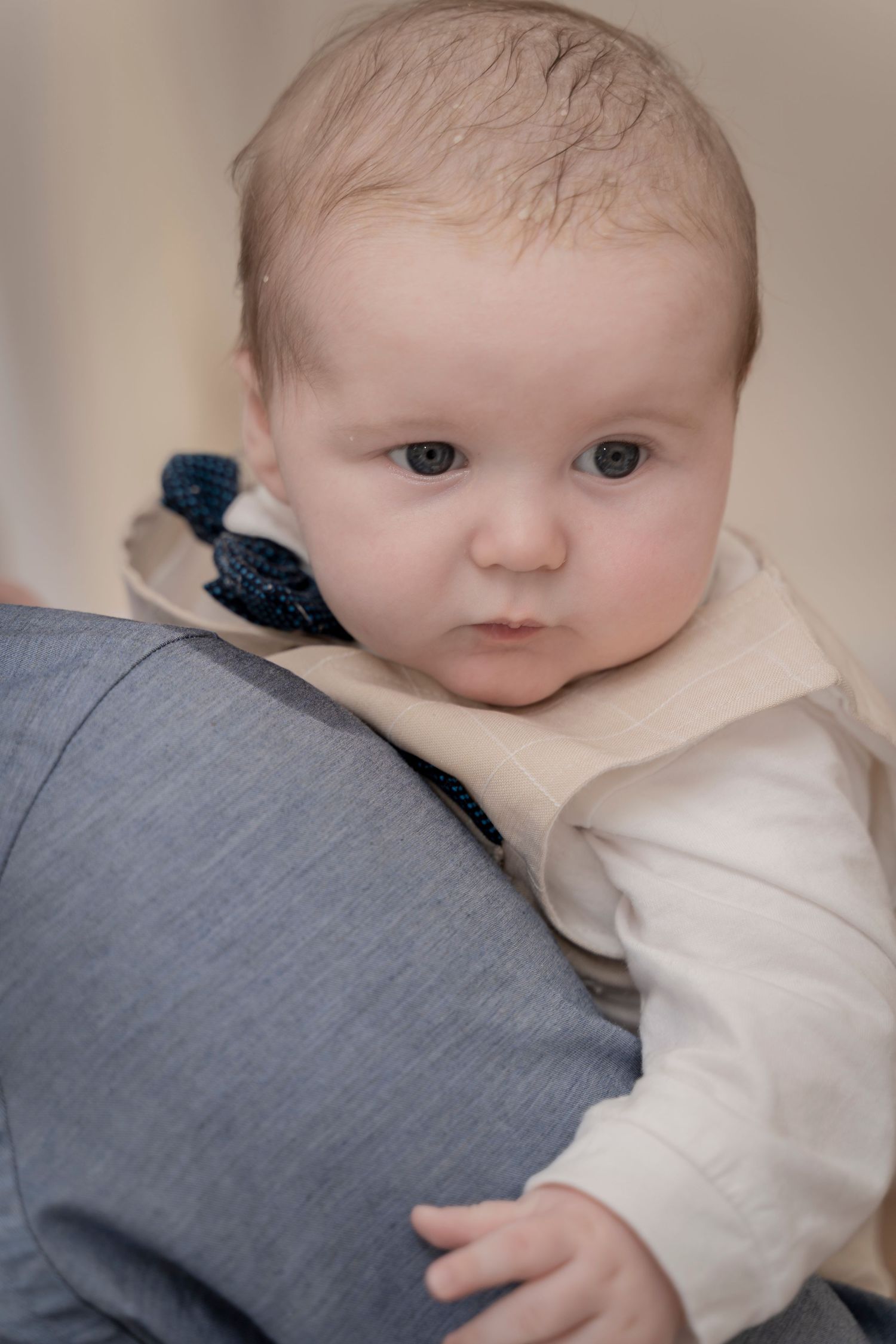 Portrait of an infant wearing cream colored clothing resting against gray pillow.