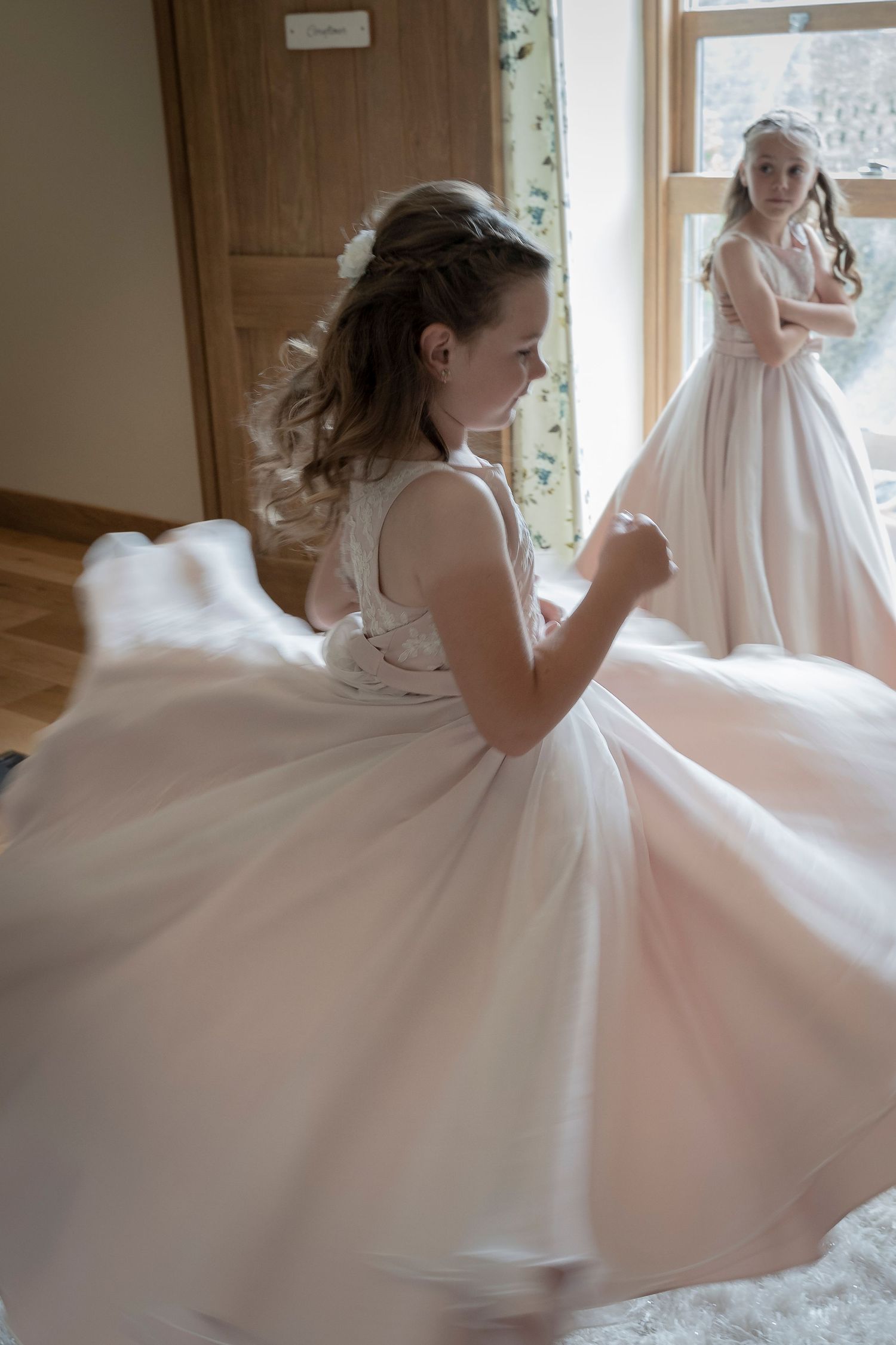 Young flower girls in flowing white dresses twirling and playing during wedding preparations.