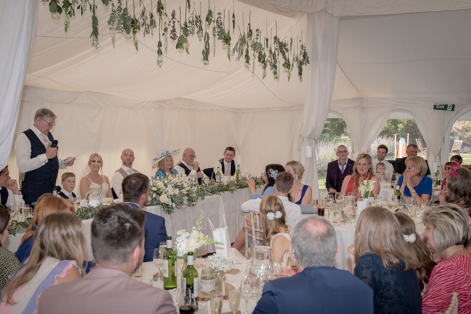 Wedding reception in white marquee tent with hanging greenery and guests seated at decorated tables. Wedding Photography Devon.