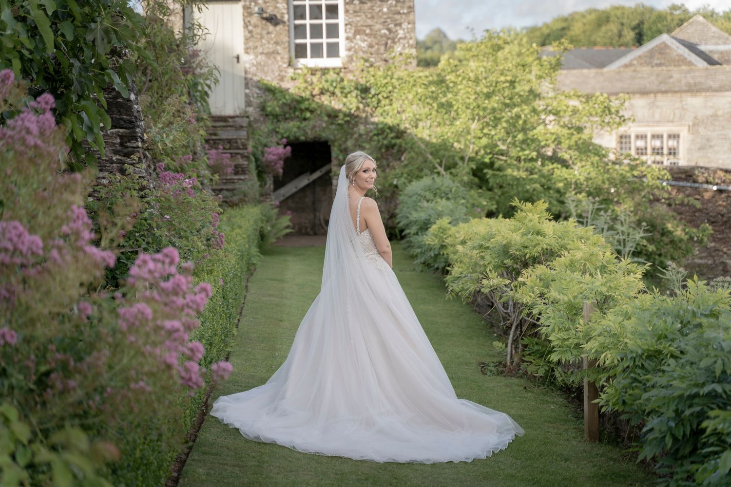Bride in flowing white gown walking through manicured garden path with stone buildings.