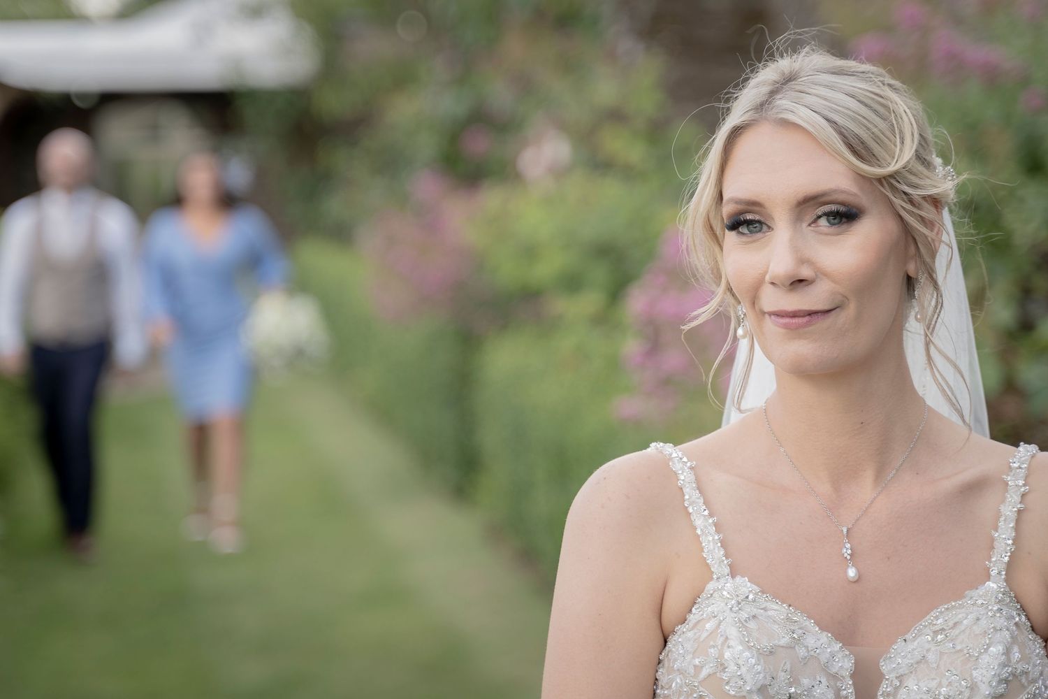 A bride in a white lace gown stands in a garden setting with soft lighting and blurred figures in background.