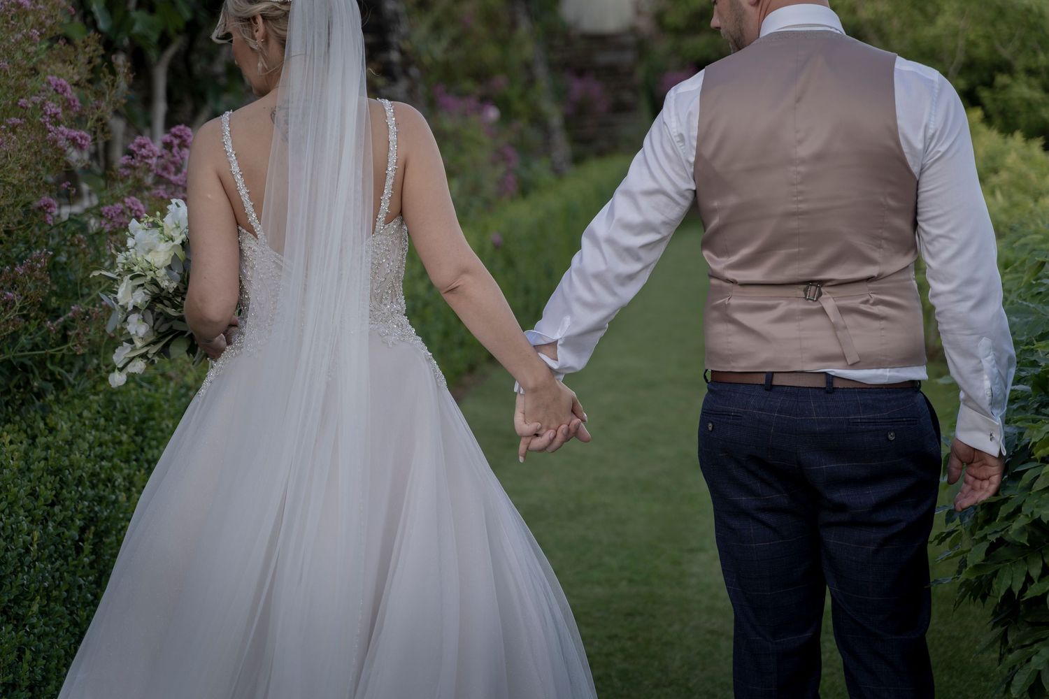 Back view of a bride and groom holding hands while walking through a lush garden pathway.