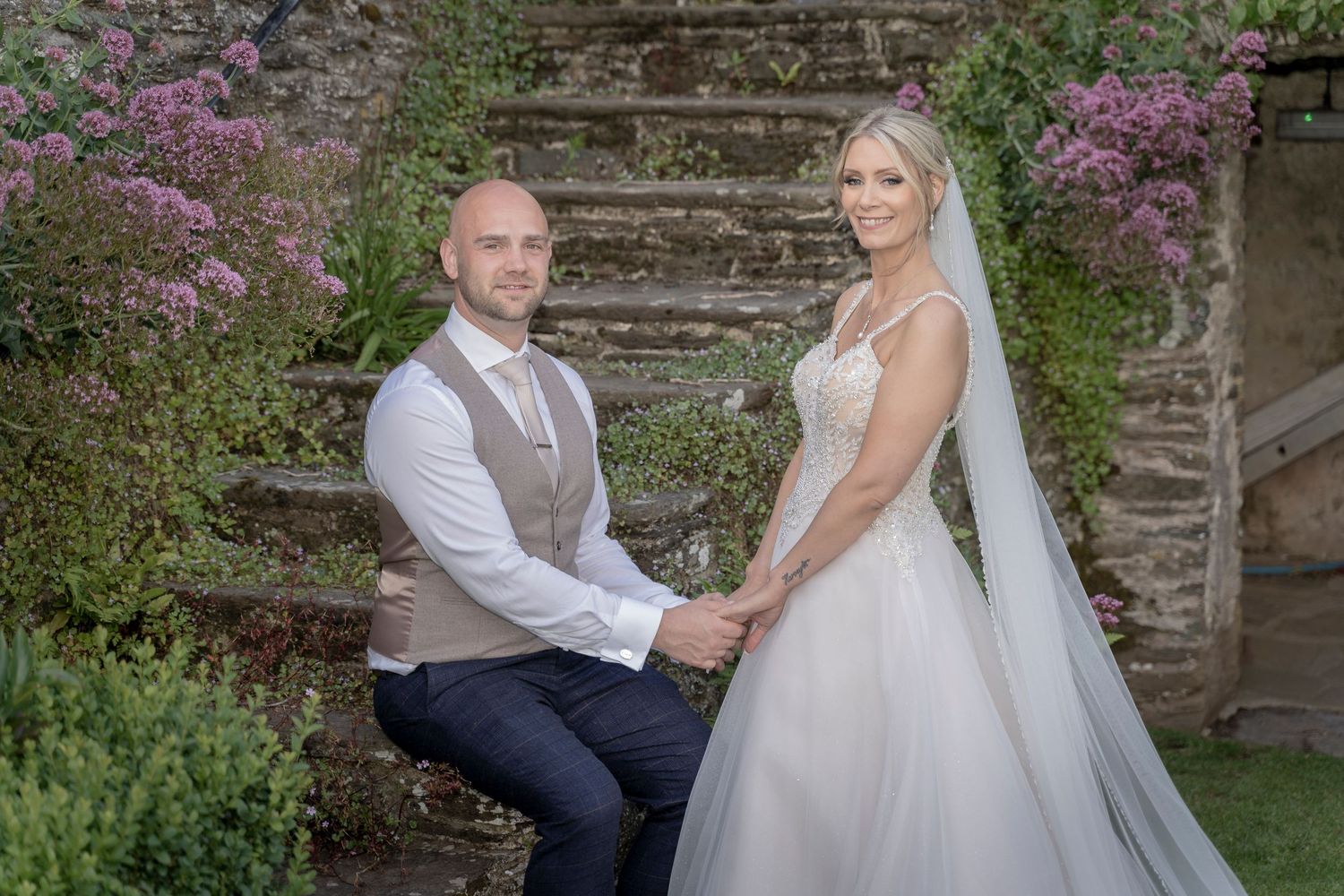 A couple in wedding attire poses on stone steps surrounded by purple flowers and greenery. Wedding reception, Devon Wedding photographer.
