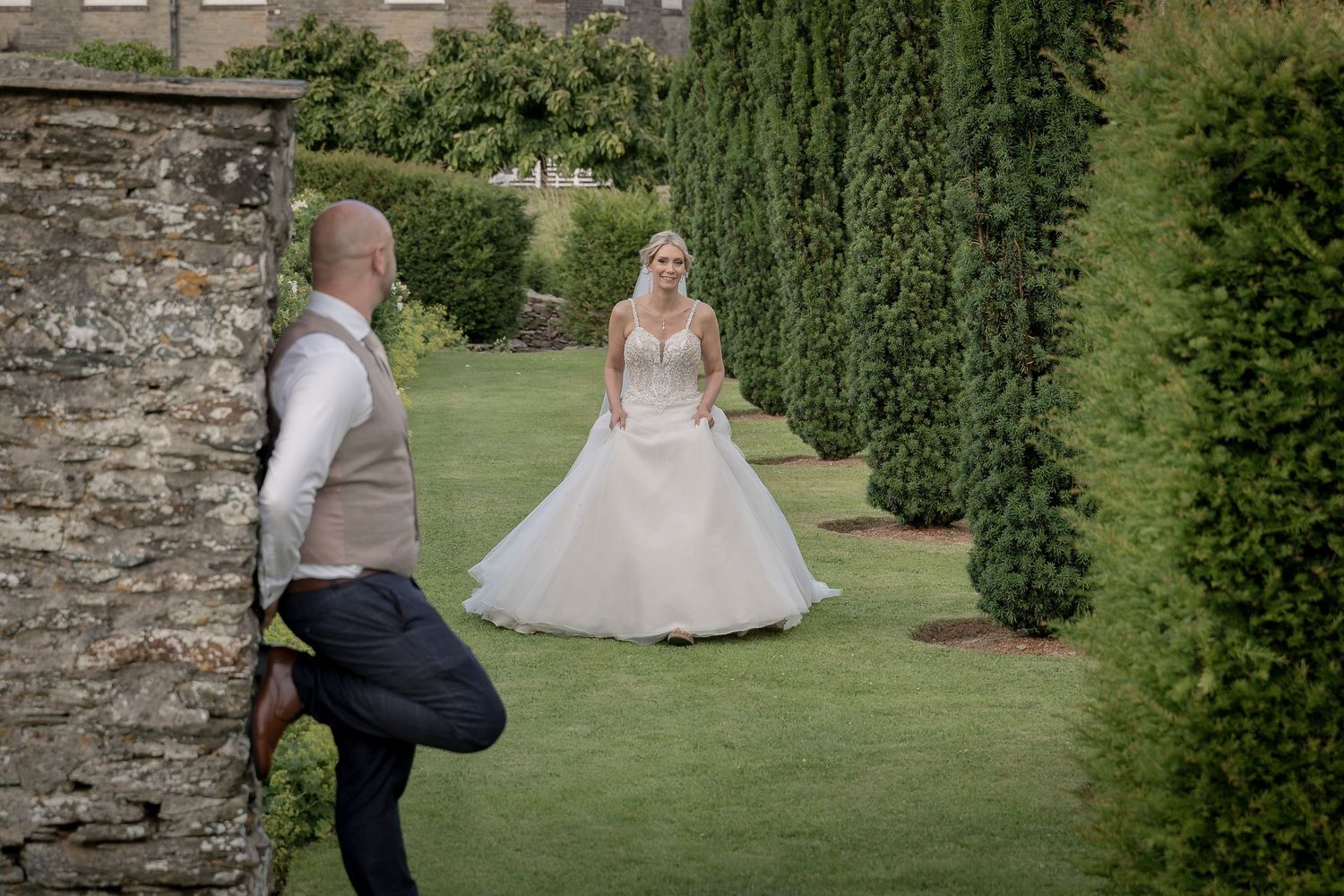 A wedding couple shares a romantic moment in a manicured garden with stone walls and cypress trees. Devon wedding photographer.