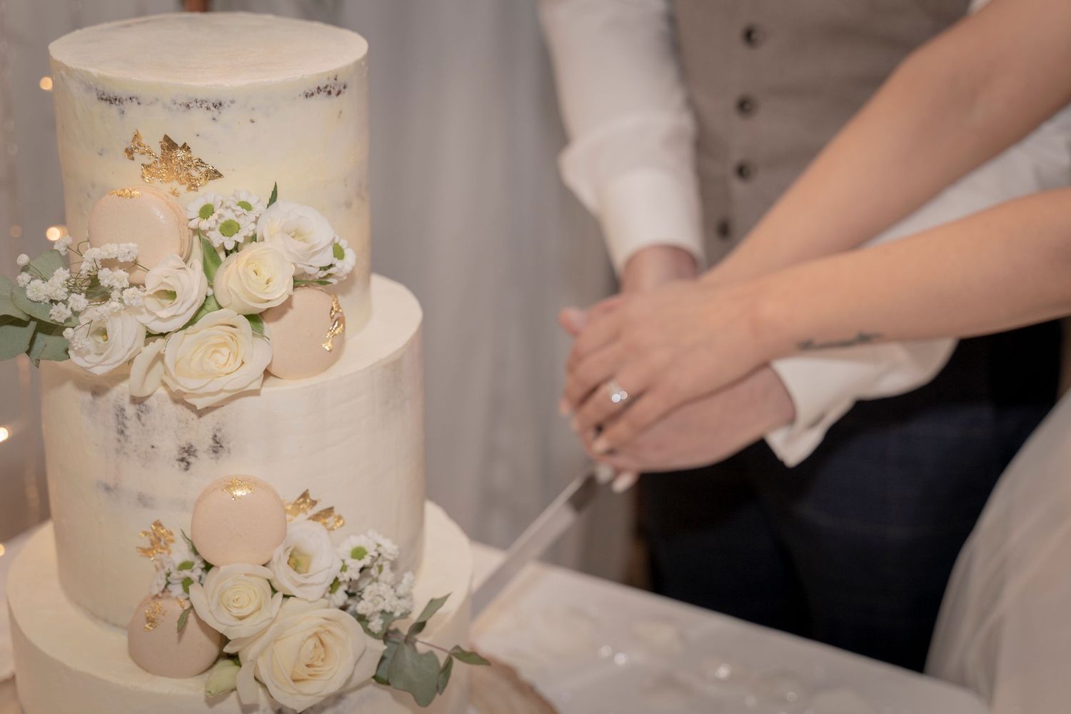 An elegant white wedding cake decorated with fresh roses and greenery being cut by newlyweds. Devon Wedding Photography.