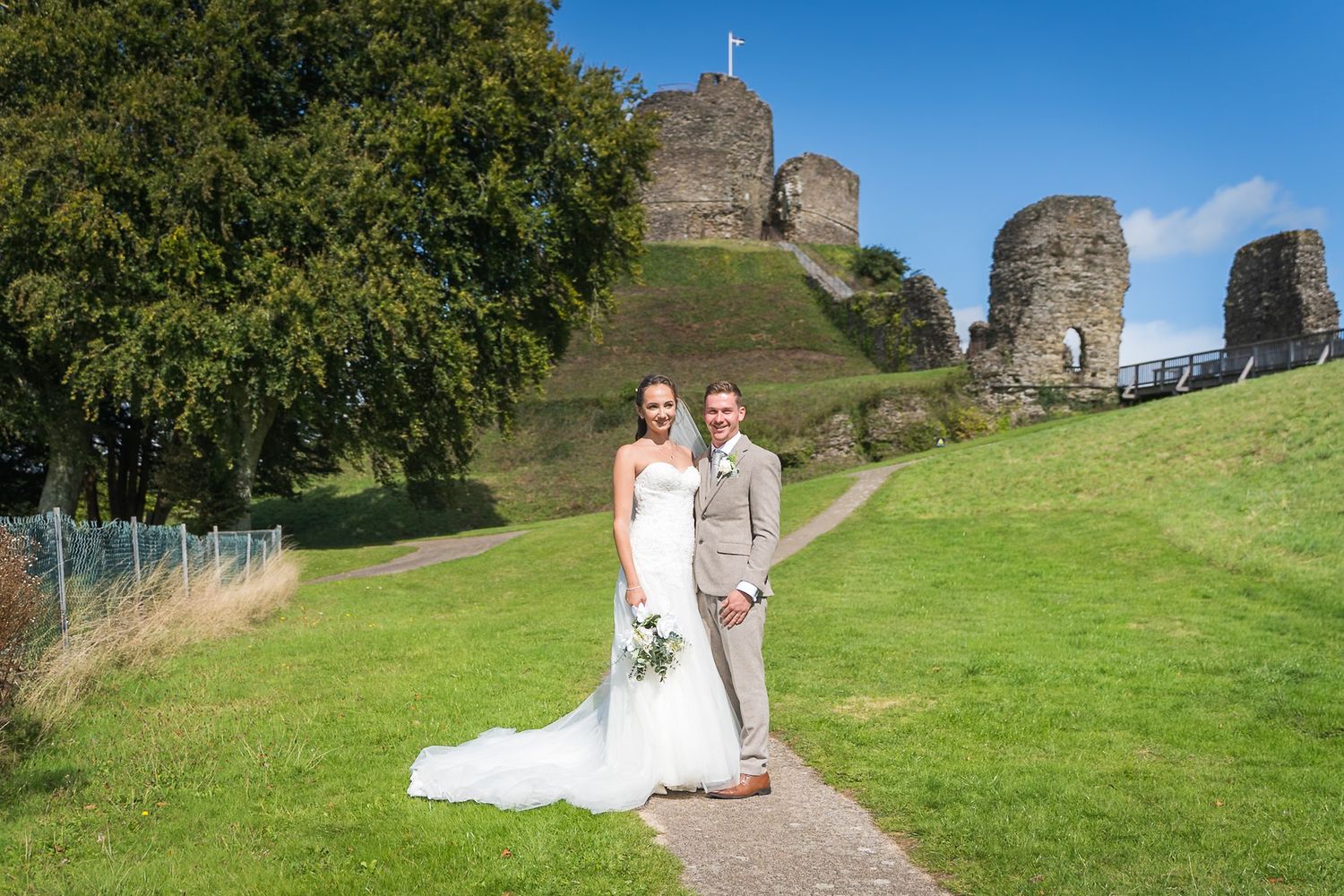 A wedding couple poses for photos on a path with ancient castle ruins and green hills in the background.