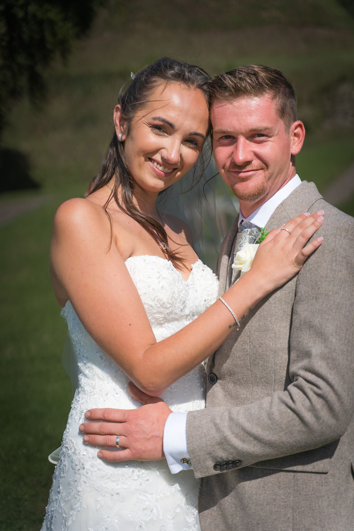 A bride in a white lace dress and groom in gray suit embrace outdoors on their wedding day.
