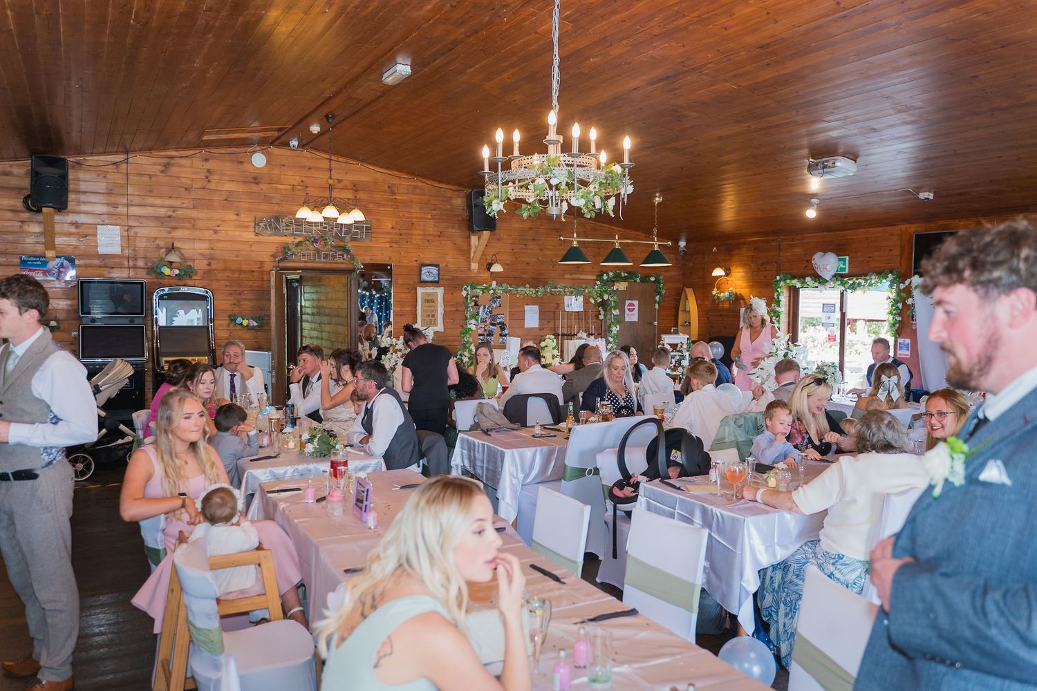 Wedding reception guests gather in a rustic venue with wooden ceiling and elegant chandelier.