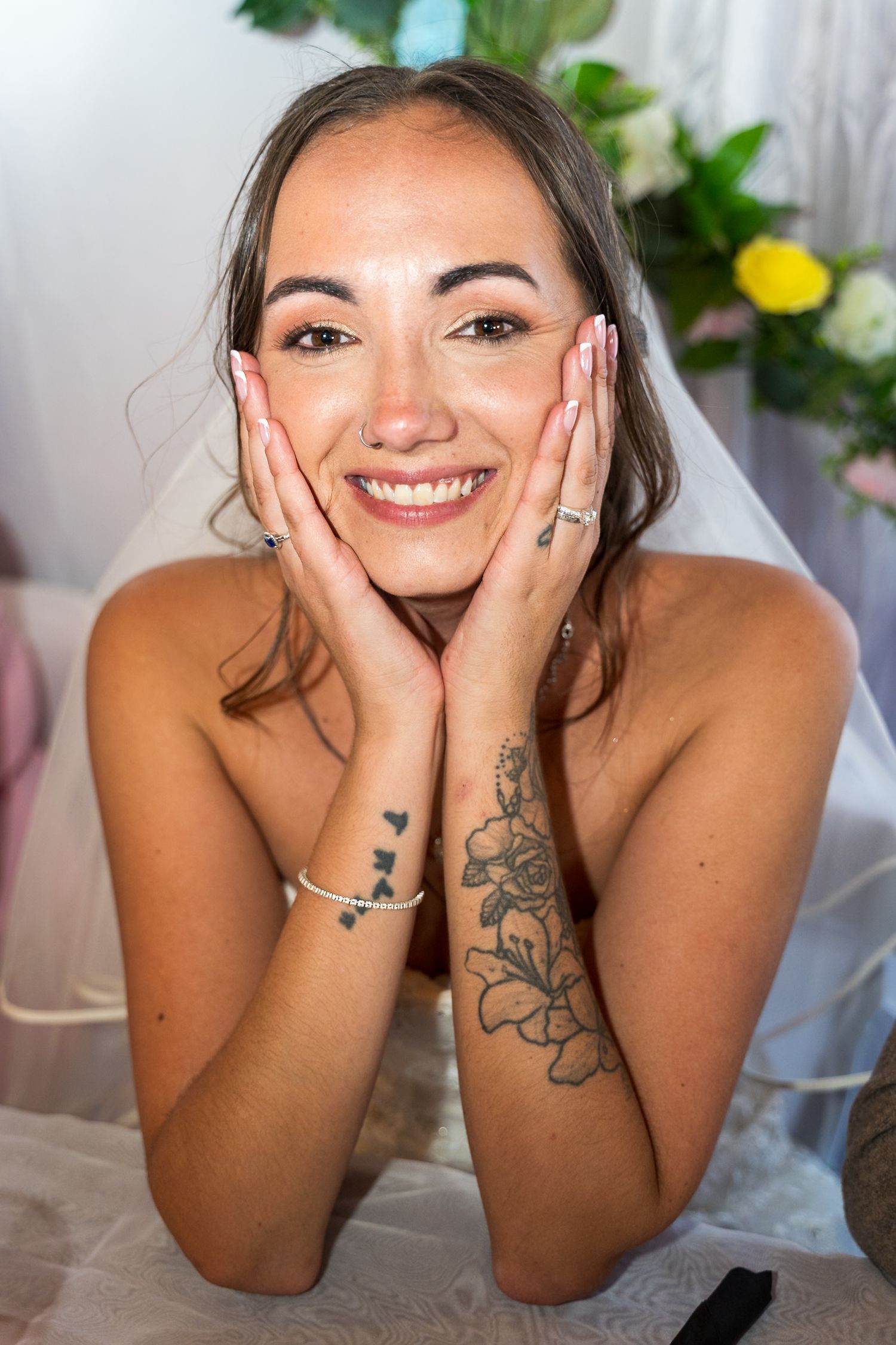 Joyful bride poses with hands on cheeks showing off wedding ring and floral tattoo.