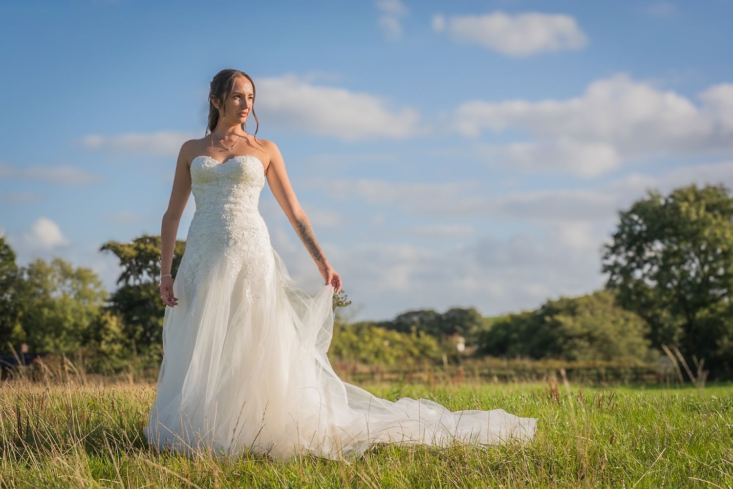A bride in a flowing white wedding dress stands in a sunlit meadow with trees and blue sky in the background.