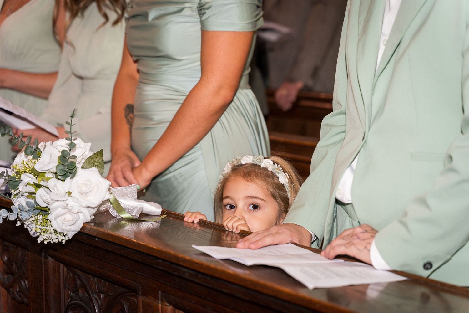A small child peeks playfully over a church pew during a wedding ceremony while surrounded by mint green attire.