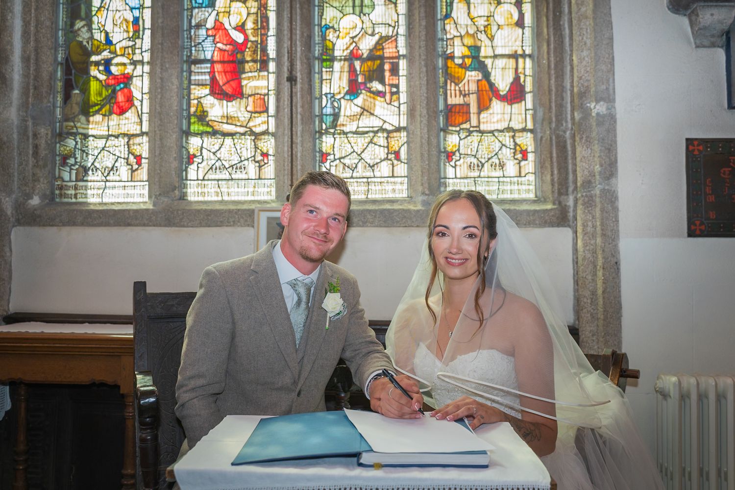 A couple signs their marriage register at a church altar beneath ornate stained glass windows depicting religious scenes.