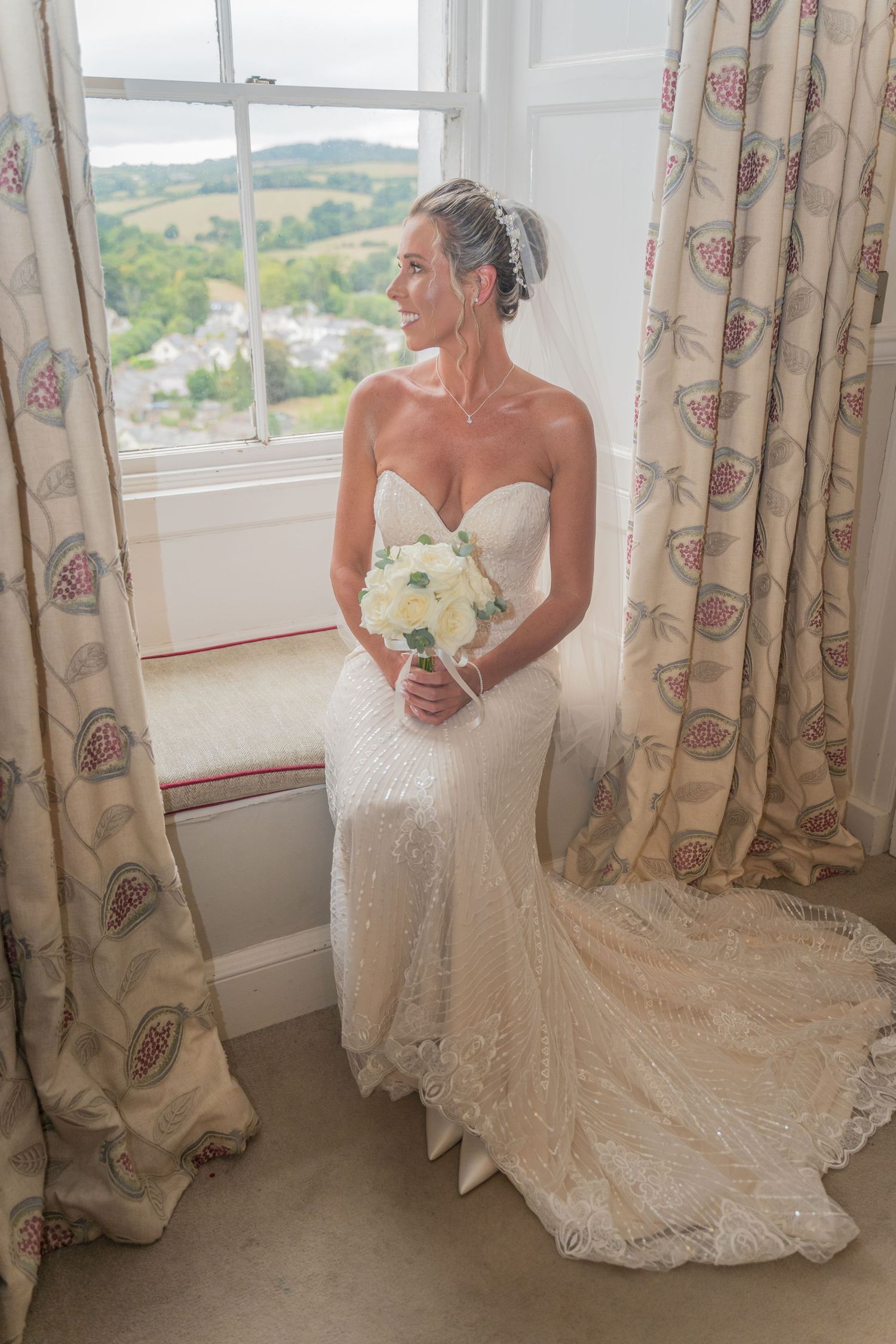 A bride in a fitted lace wedding gown with a sweetheart neckline holds a white bouquet while standing by a window.