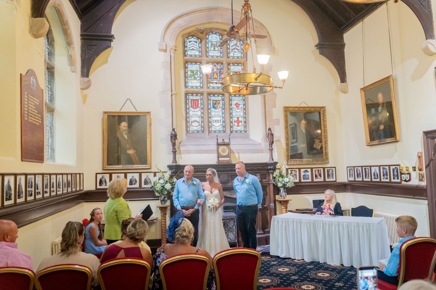 People gather in an elegant church hall with stained glass windows and portrait-lined walls for a formal event.