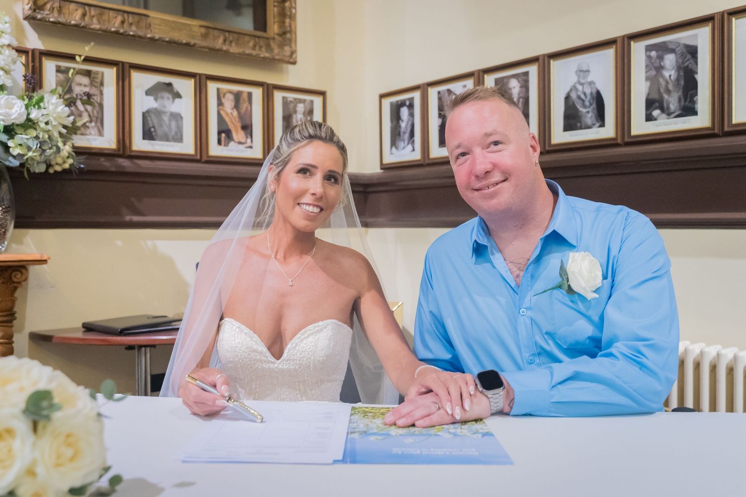 Couple in formal wedding attire signing documents together at a wooden table during a ceremony.