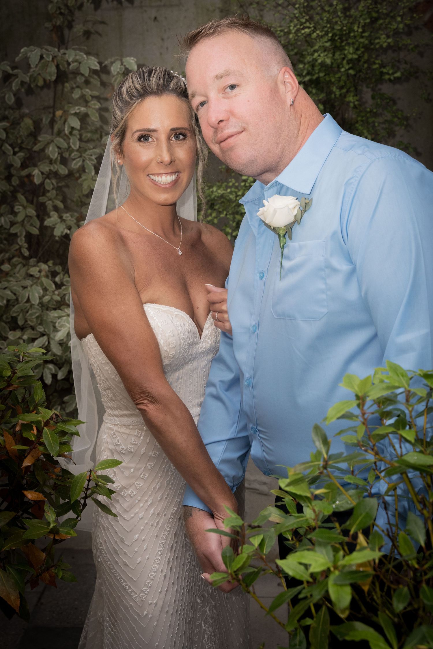 Couple in wedding attire poses together against lush greenery backdrop in romantic outdoor setting.