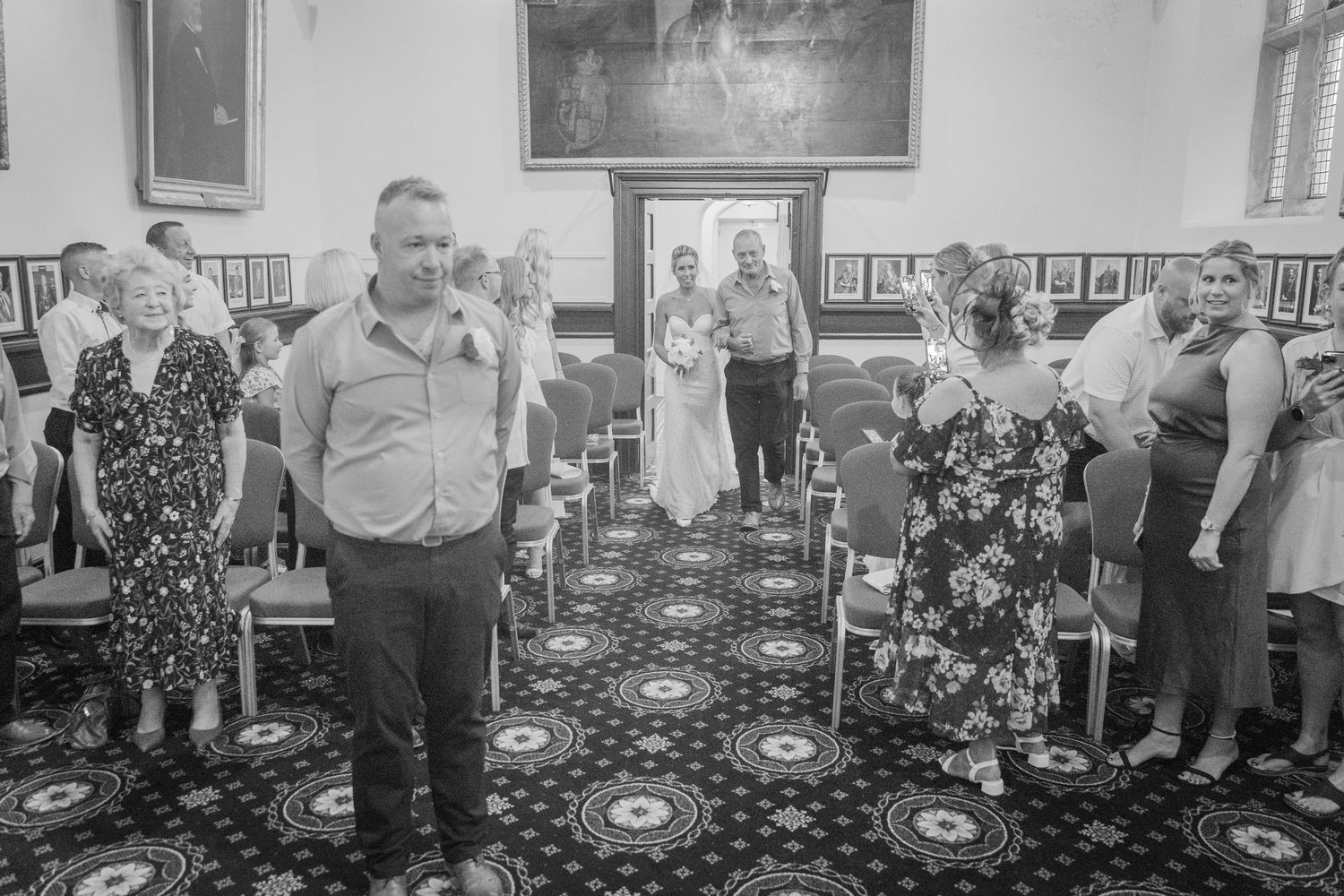 A black and white photograph of people gathered in an ornate room with patterned carpeting and framed artwork on the walls.