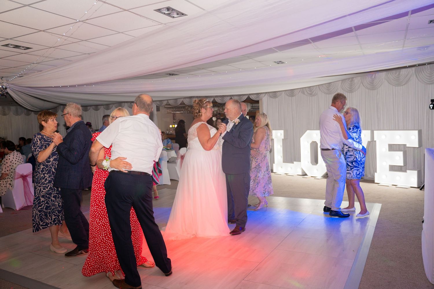 Couples dancing together in a spacious ballroom with white ceiling tiles and colorful mood lighting.
