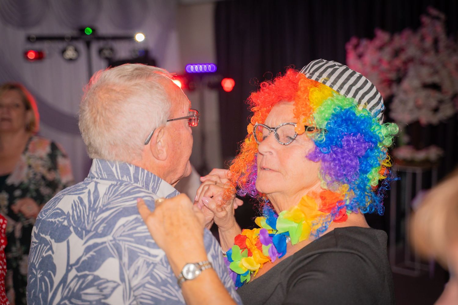 Two people share a fun moment at a party, with one wearing a colorful rainbow wig and the other in a light patterned shirt.