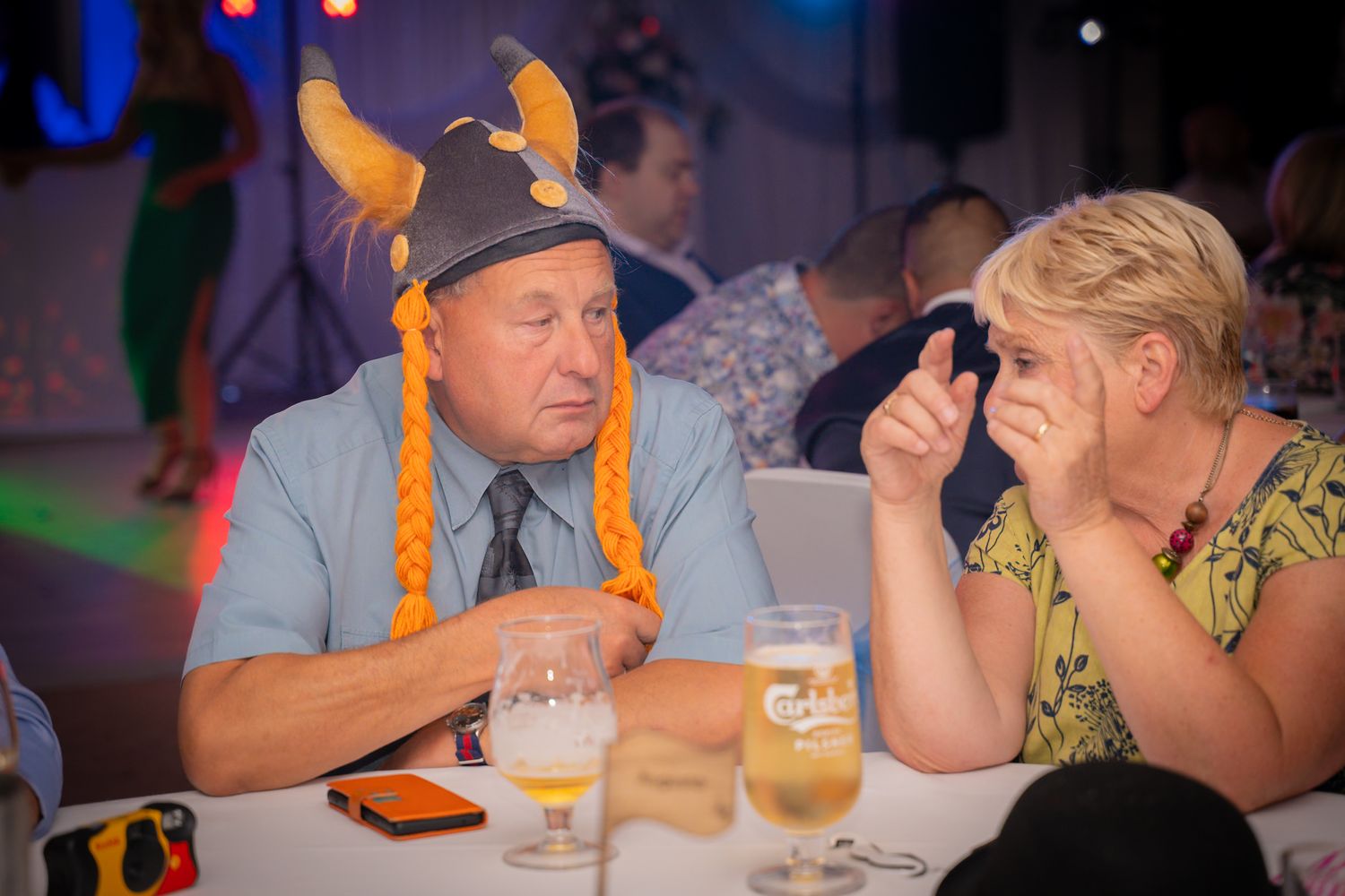Two guests at a party sit at a table, with one wearing a novelty orange horned hat.