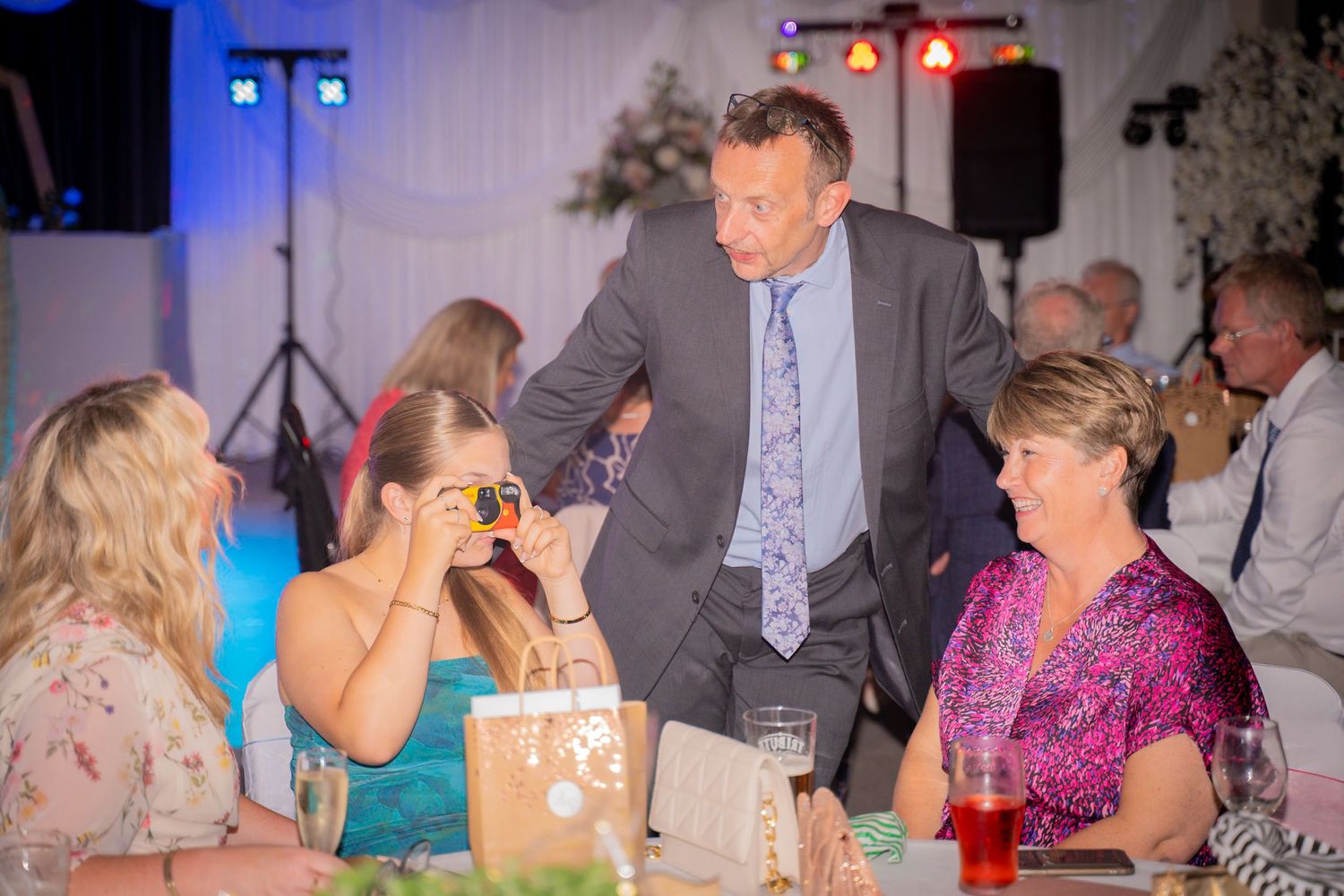 Wedding reception guests share laughter at decorated table with blue mood lighting.