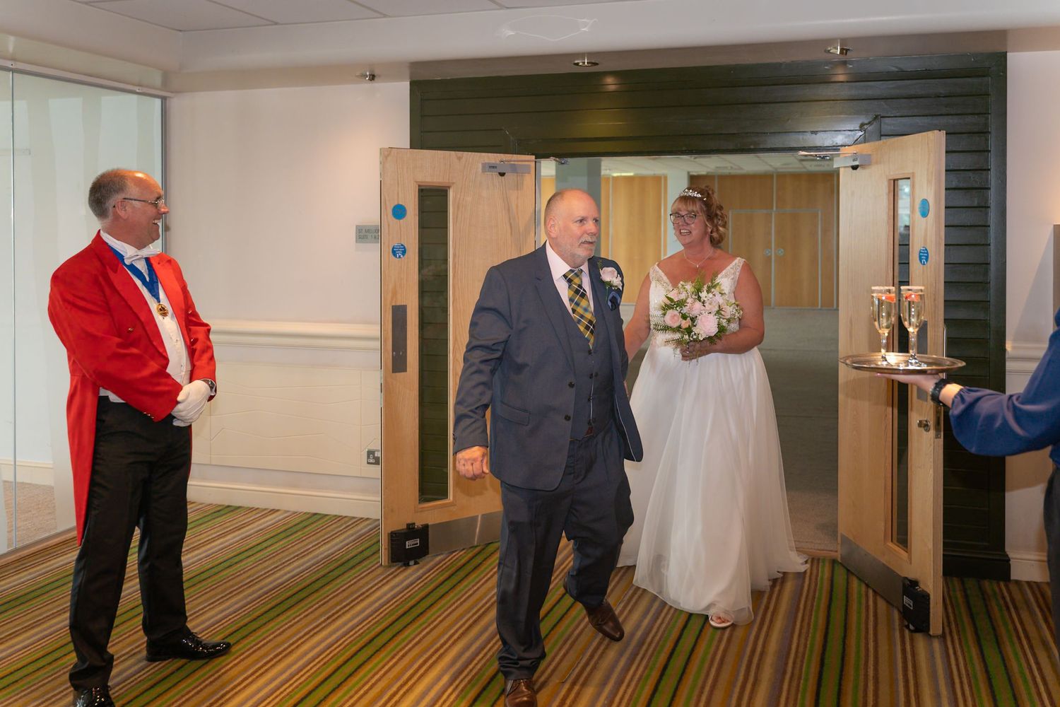 A couple in wedding attire enters through glass doors at a venue while a person in a red jacket stands nearby.