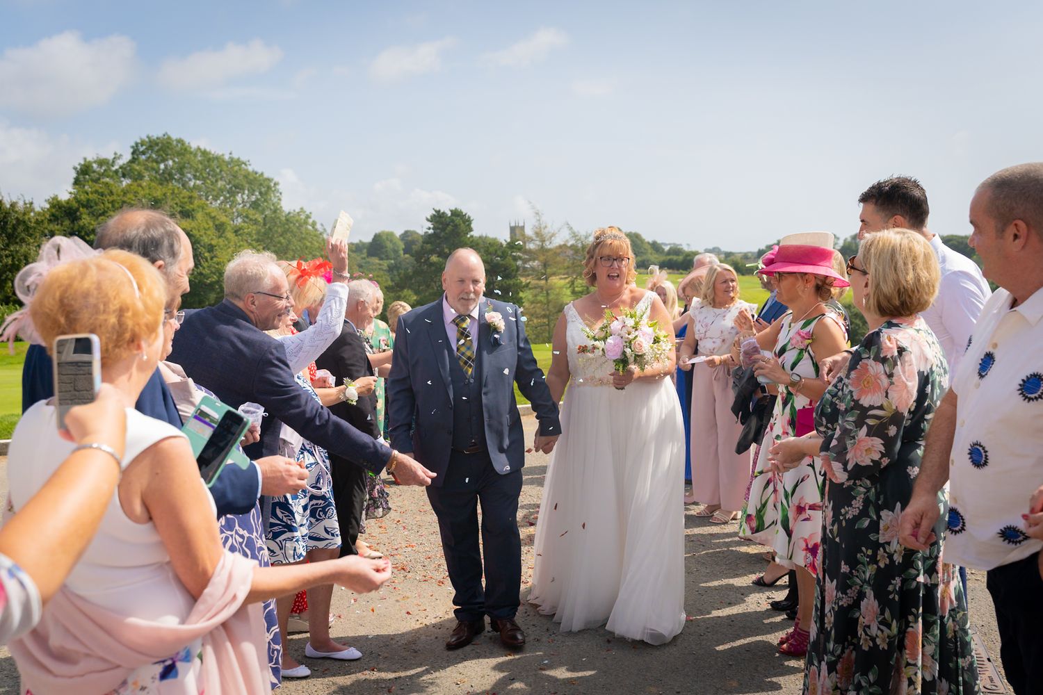 Wedding guests gather outdoors on a sunny day to celebrate and take photos at a scenic venue.