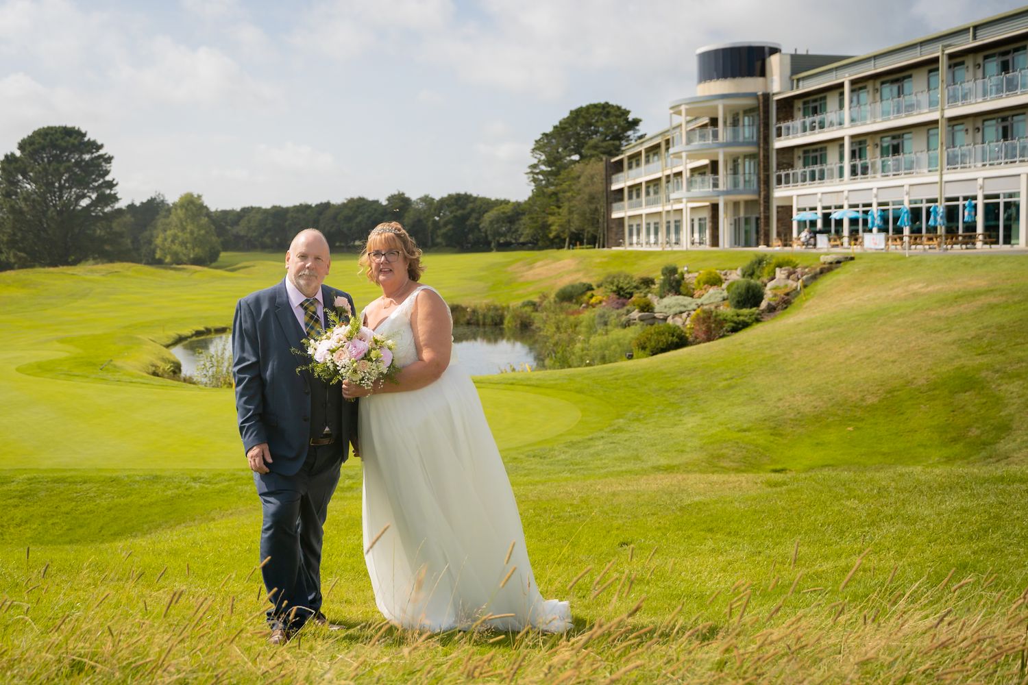 Wedding couple stands together on a lush green golf course with modern hotel building in background.