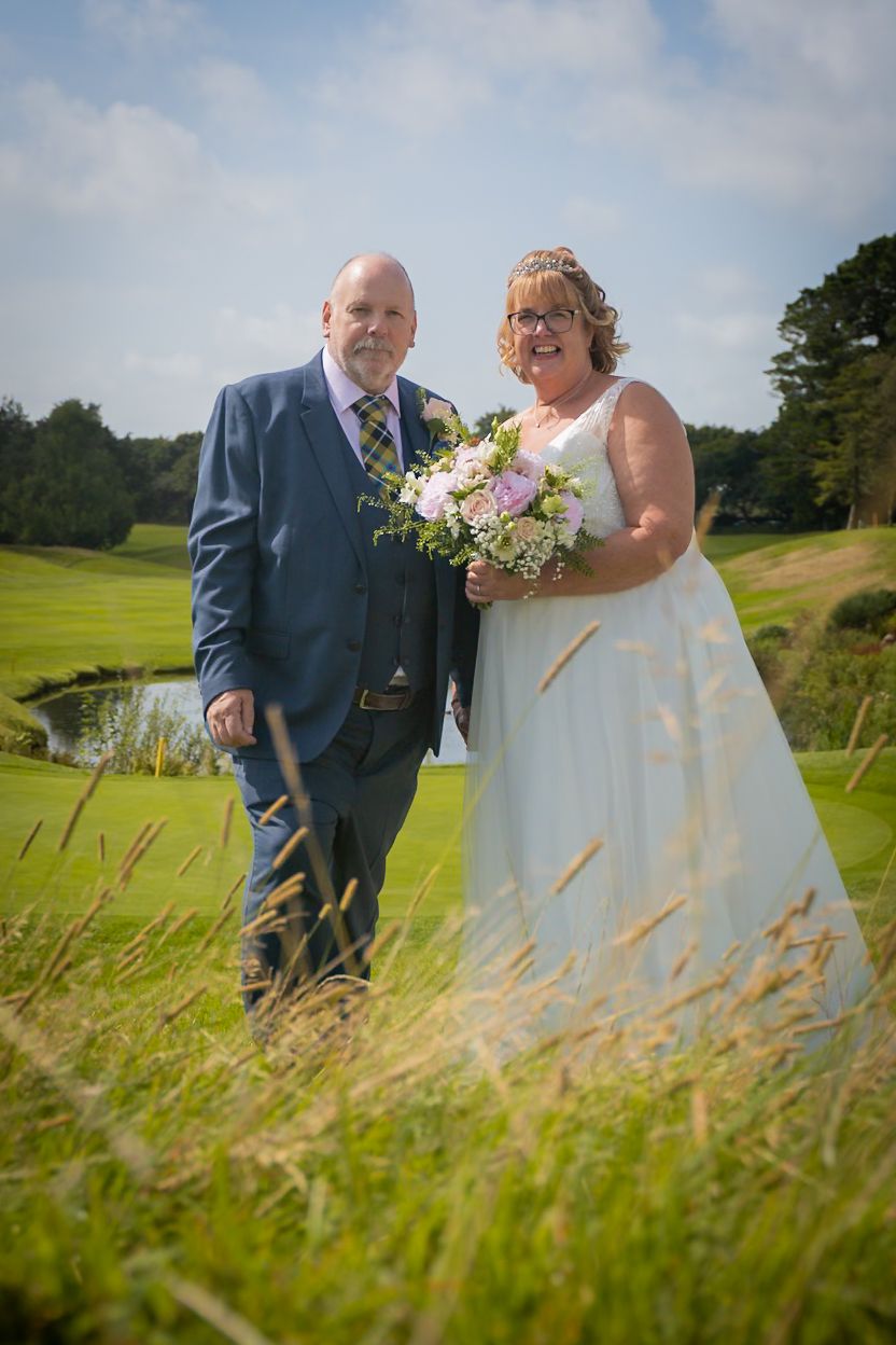 A wedding couple stands together on a lush golf course with tall grass in the foreground and trees in the background.