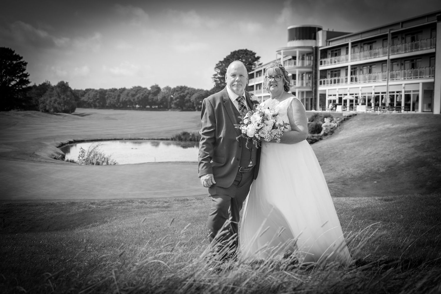 Dramatic black and white wedding portrait on golf course with modern building and stormy sky.