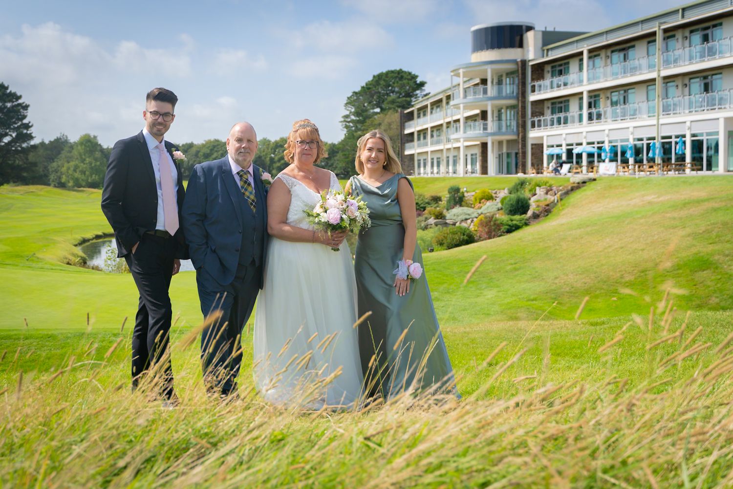 Wedding party stands on green hillside overlooking coastal resort hotel with panoramic ocean views.
