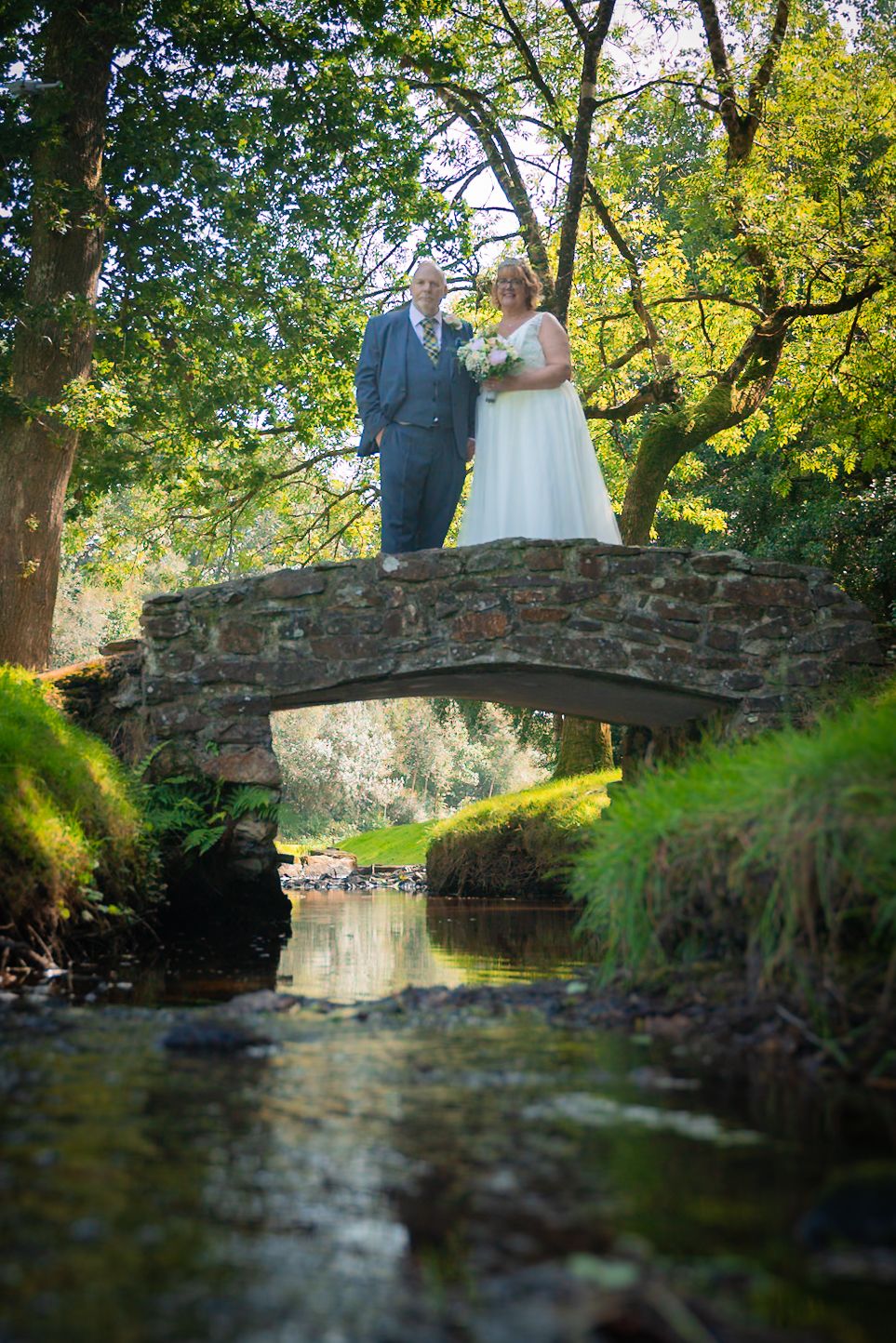 Romantic wedding photo on rustic stone bridge over stream with sunlit trees in background.