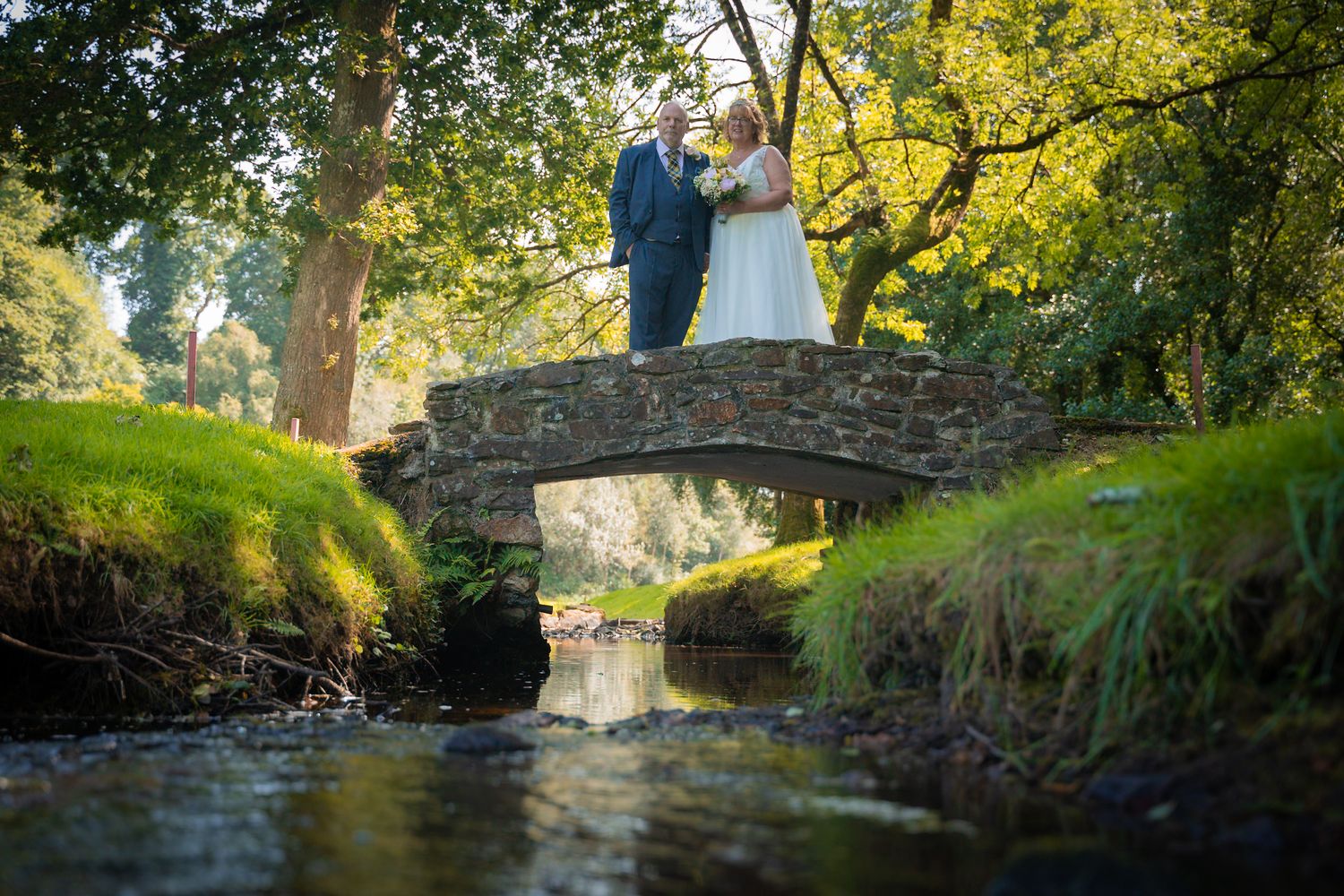 Wedding party poses on a rustic stone bridge surrounded by lush green trees and foliage.