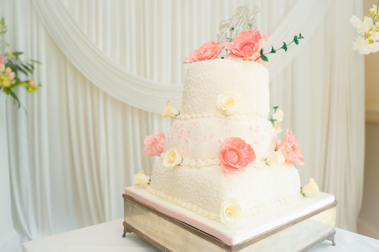 Elegant white wedding cake decorated with pink roses and floral accents displayed against draped fabric backdrop.