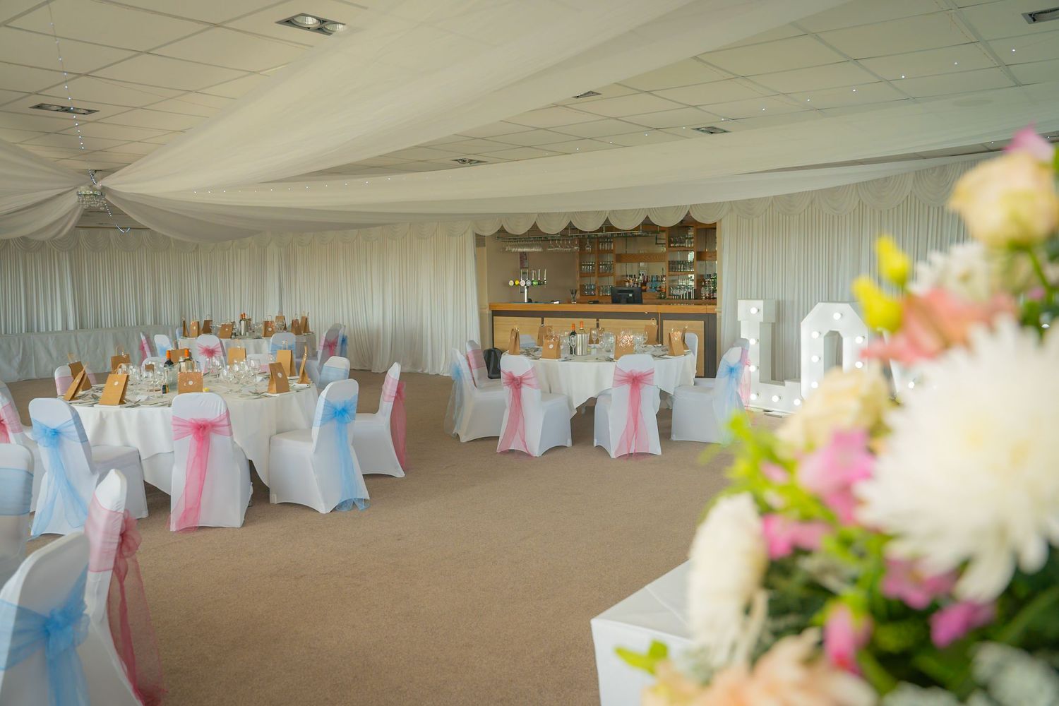 Elegant wedding reception venue with white draping, crystal chandeliers, and pink and blue accent lighting.