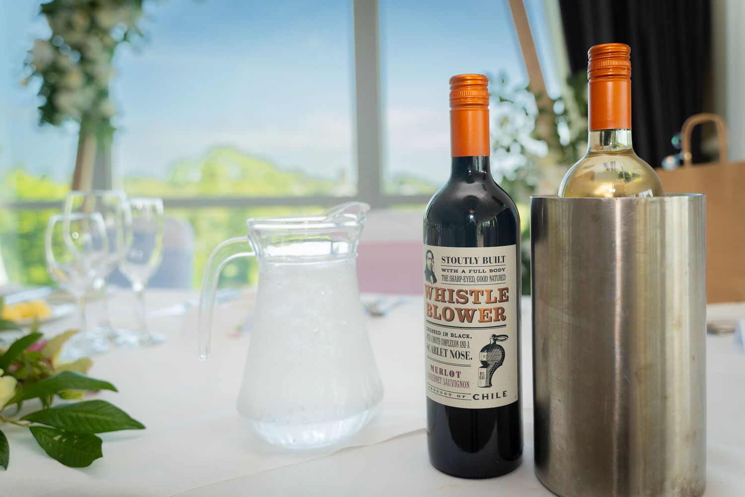 Wine bottles and glasses arranged on a bright windowsill with a stainless steel wine chiller and natural light streaming in.