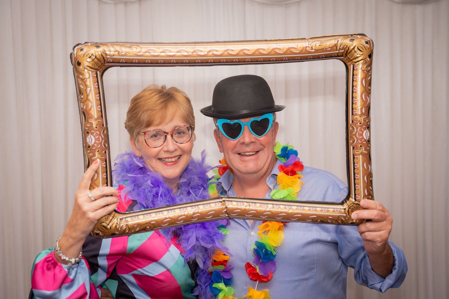 People holding a decorative gold picture frame pose with party accessories and colorful leis at an event.