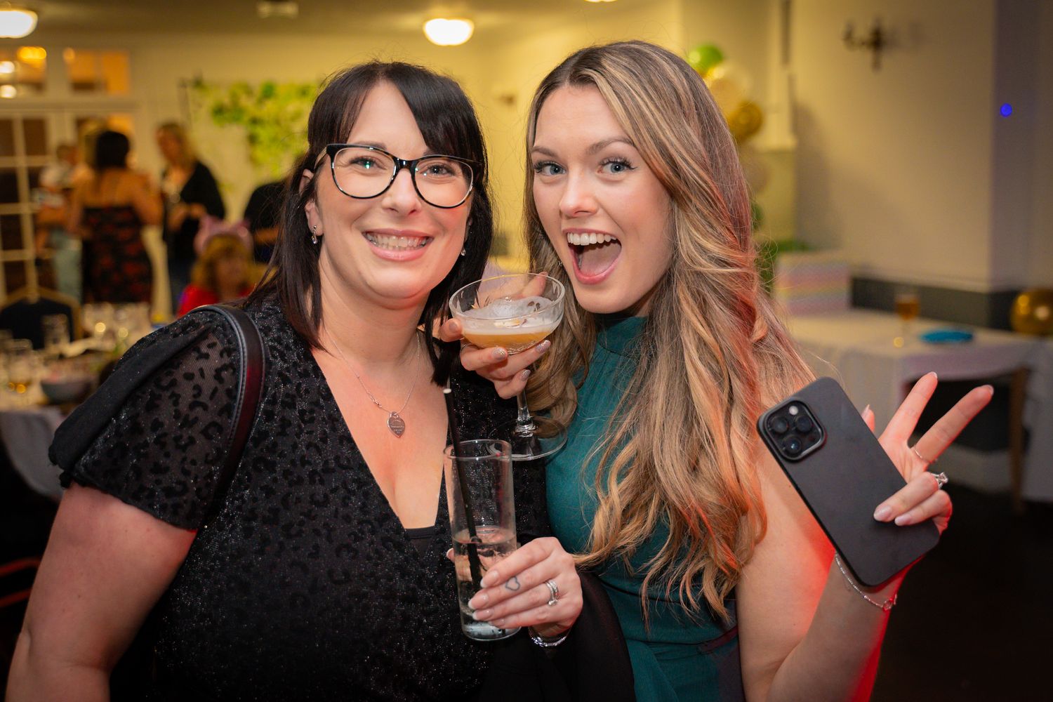 Two friends enjoying drinks and laughing together at a social gathering in an indoor setting.