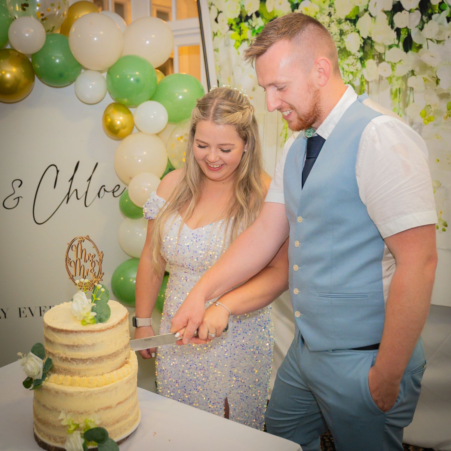 A couple in formal attire cuts into a white and gold wedding cake adorned with floral decorations at their celebration.