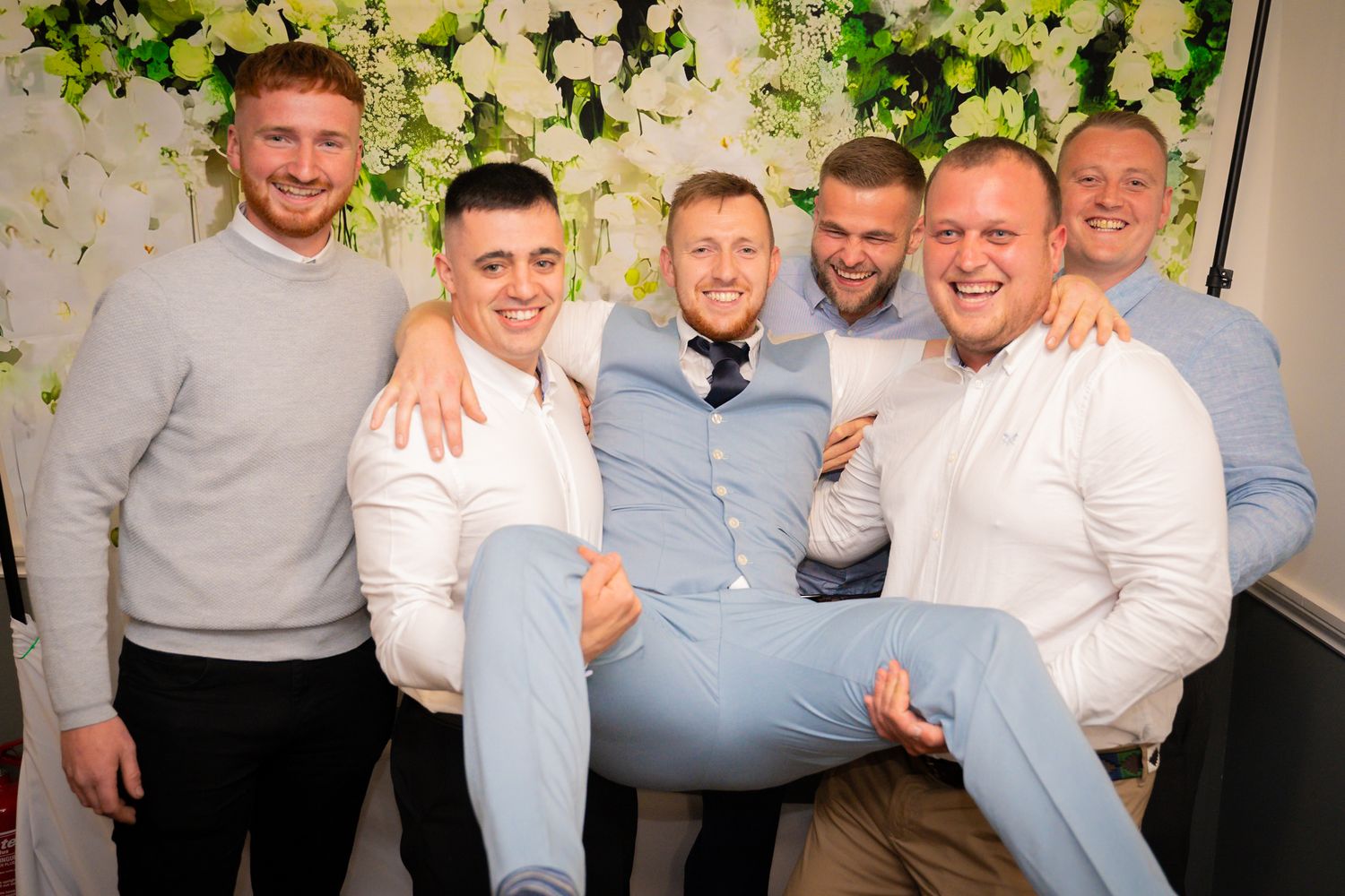 A group in matching white and blue outfits poses together against a floral backdrop at a formal event.