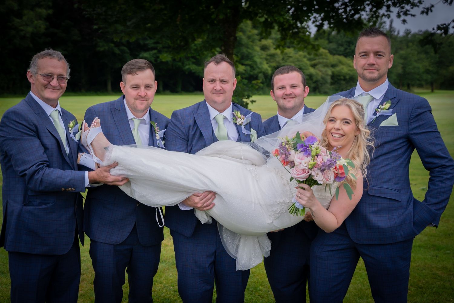 Wedding party in navy suits lifts and carries bride in white dress during outdoor photo session.
