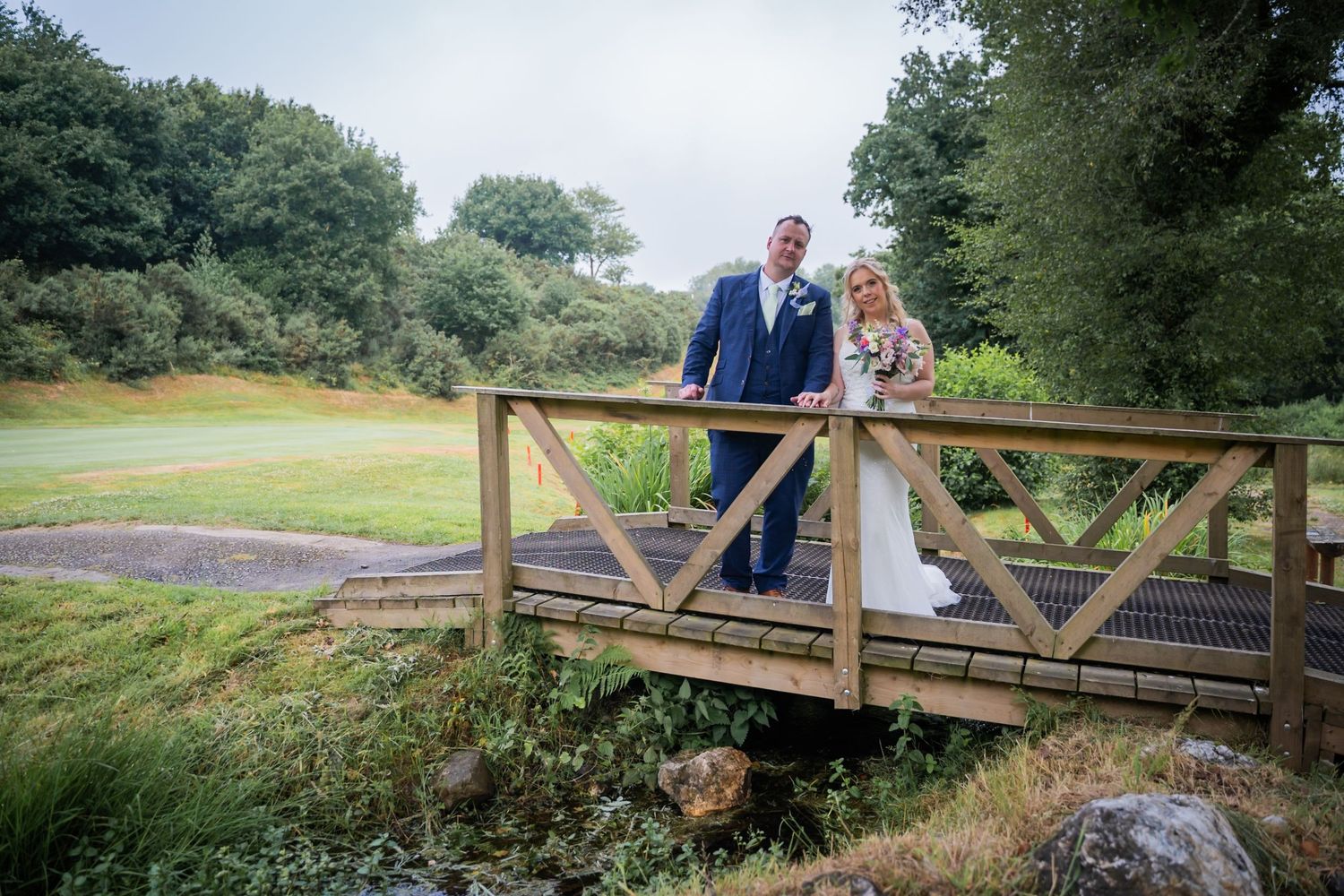 Couple standing on rustic wooden bridge overlooking scenic pond and greenery in romantic outdoor setting.